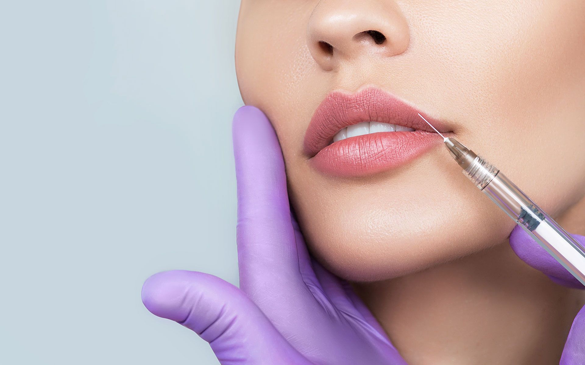 Lips being injected with a syringe, held by purple-gloved hand. Light pink lips and pale skin.