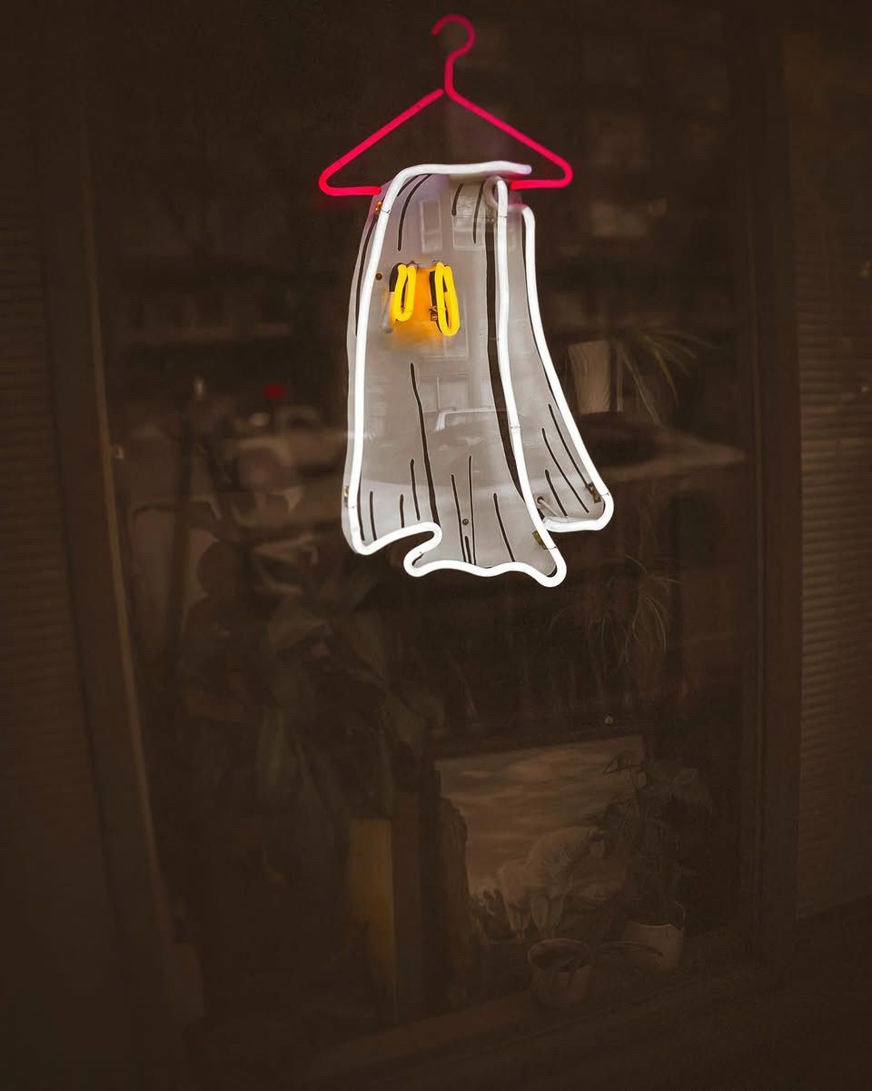 Neon ghost outline on a hanger with yellow eyes against a blurry indoor background; art by Yale Wolf. 