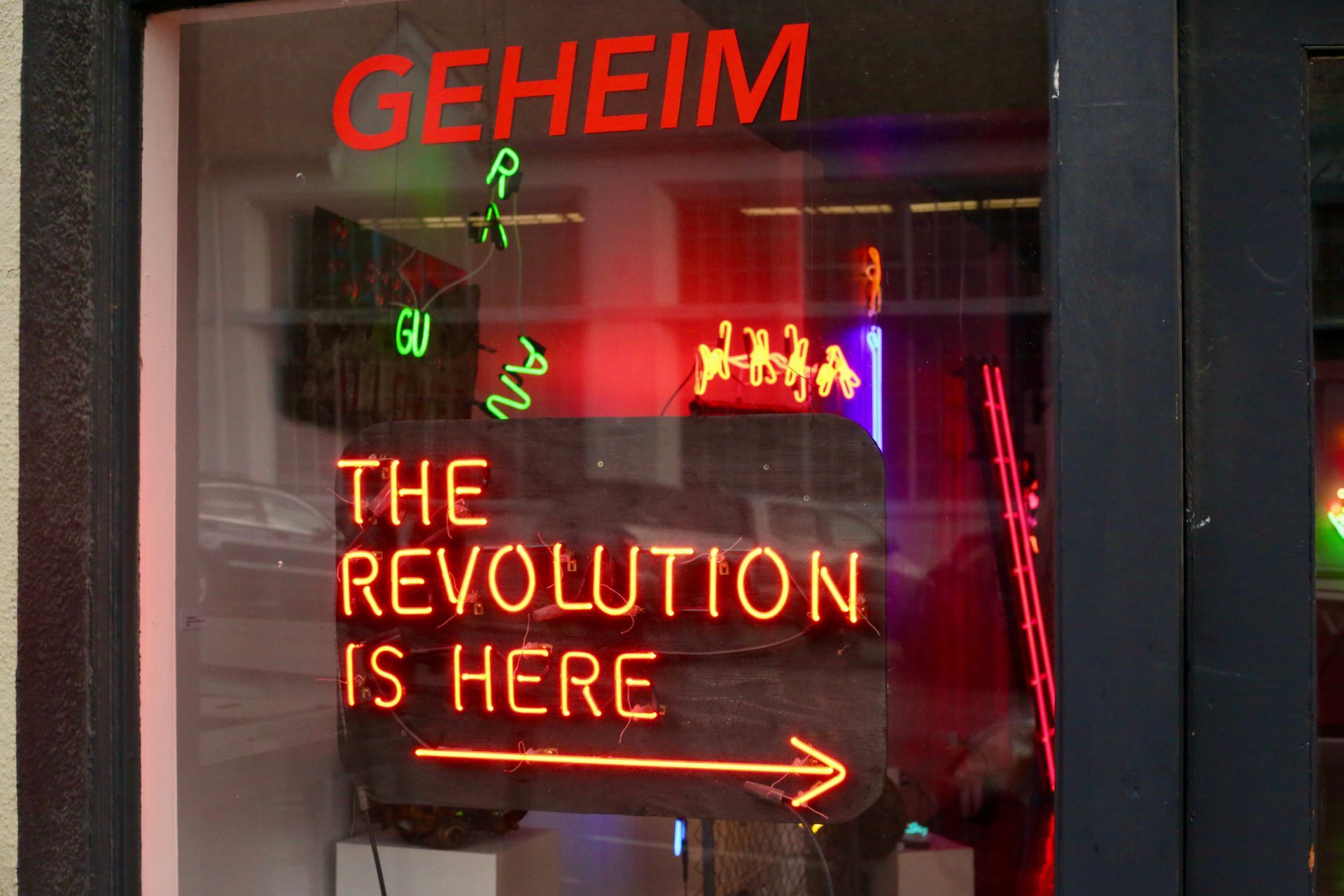 Neon sign: "GEHEIM THE REVOLUTION IS HERE" with arrow, red and green lights in a store window; by Yale Wolf.