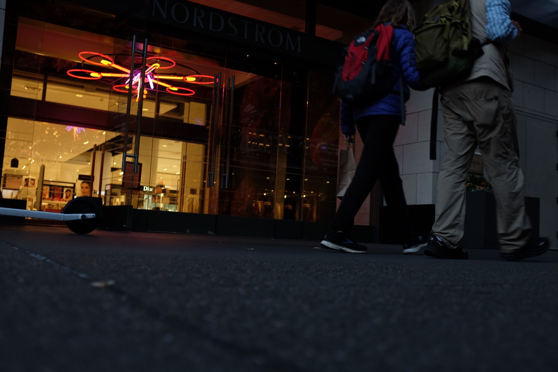 People walk past a Nordstrom storefront at night; neon art display visible; by Yale Wolf. 