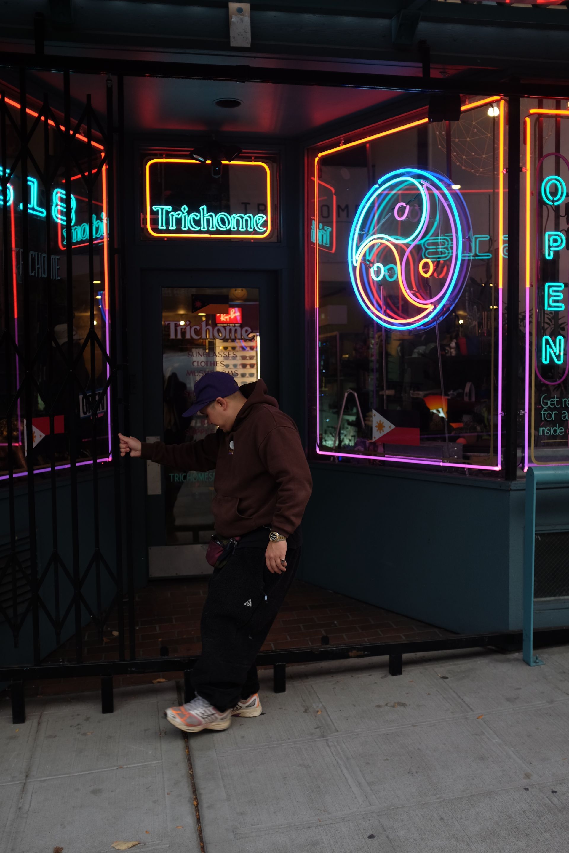 Person wearing a brown hoodie opens a metal gate outside a shop with neon signs, including a yin-yang symbol; by Yale Wolf. 