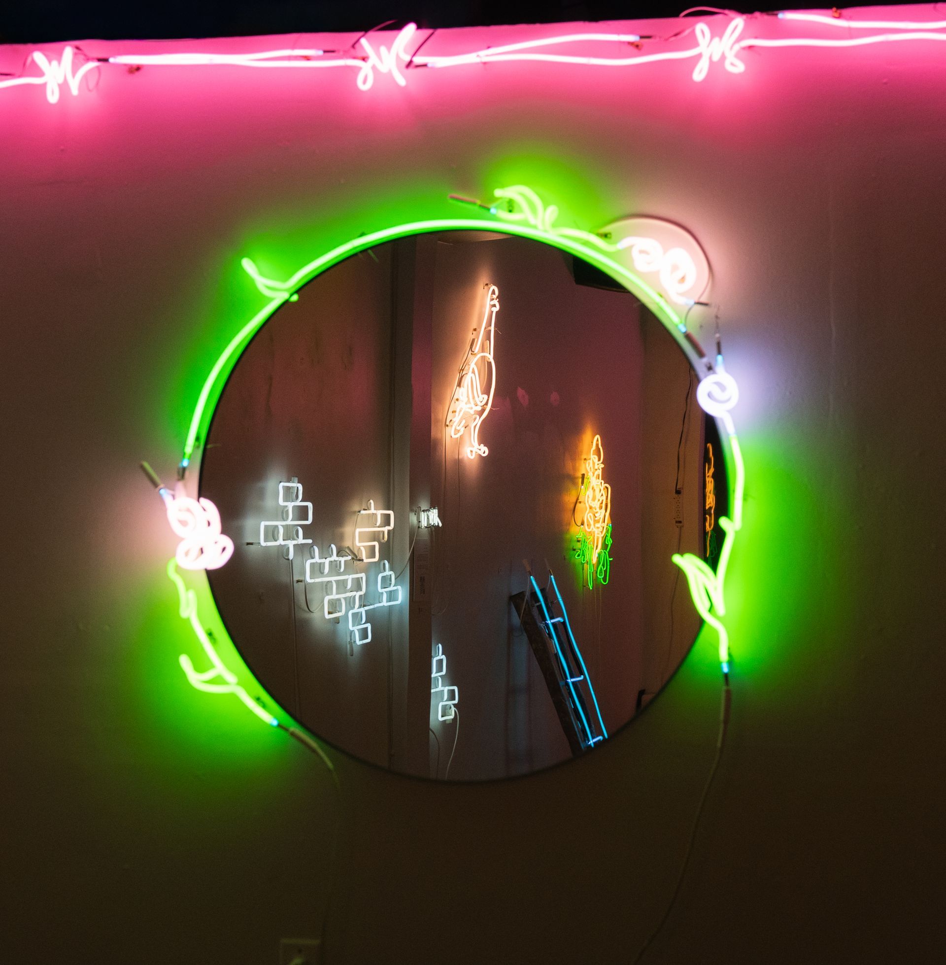 Round mirror with neon green floral outline; reflections of neon lights in the mirror. Pink neon above; art by Yale Wolf.