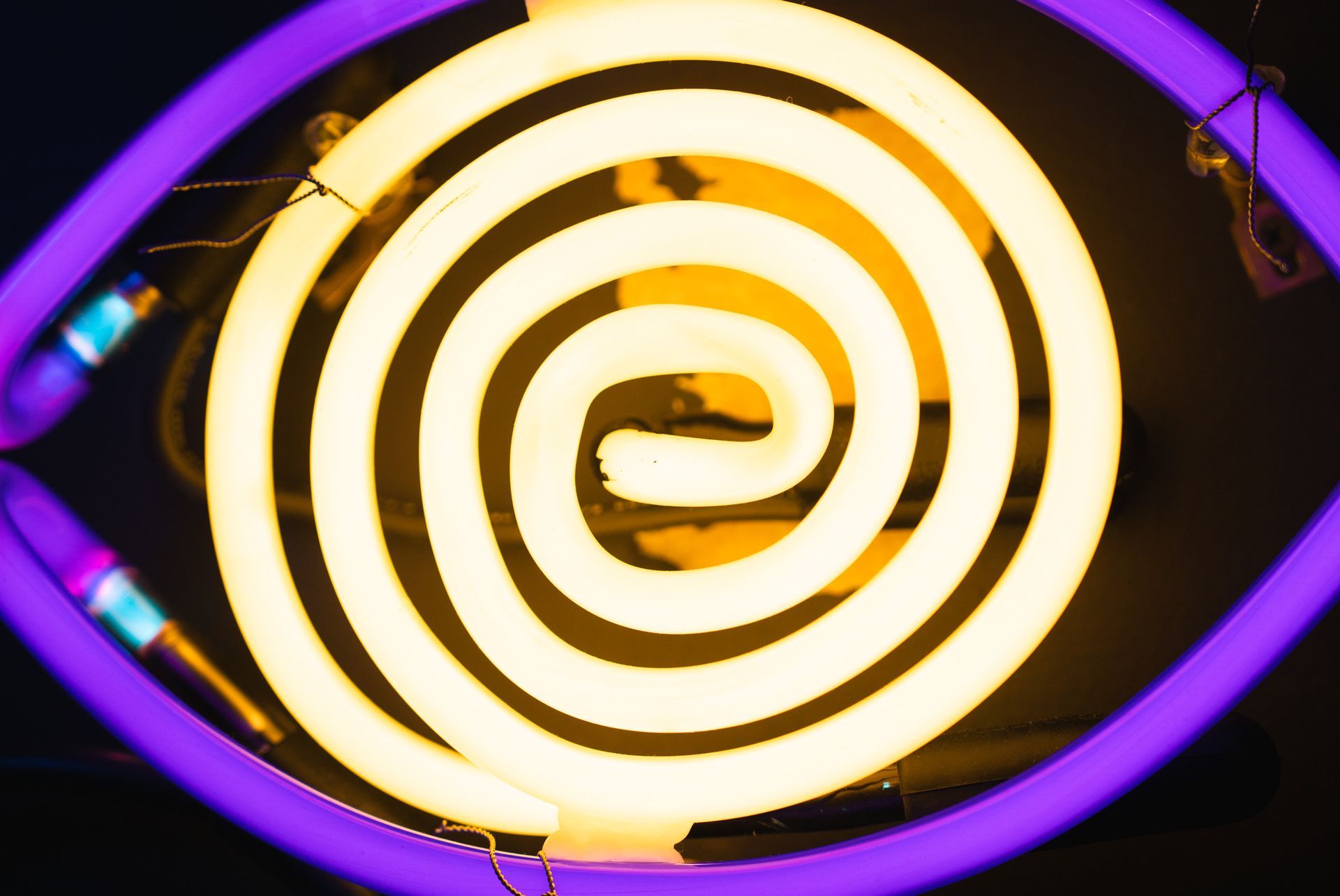 Neon eye sign with purple outline and yellow spiral iris against black; art by Yale Wolf.