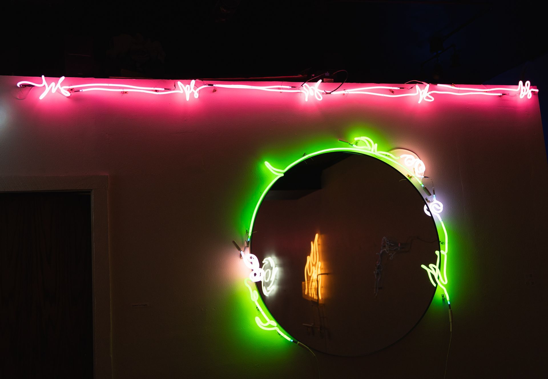 Round mirror with neon green floral outline; reflections of neon lights in the mirror. Pink neon above; art by Yale Wolf.