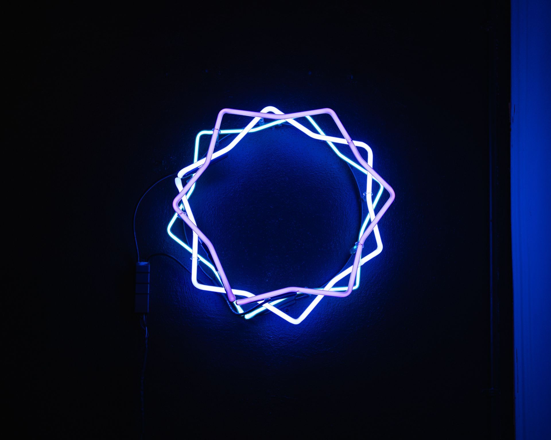 Neon blue and purple geometric star shape on a black wall; art by Yale Wolf.