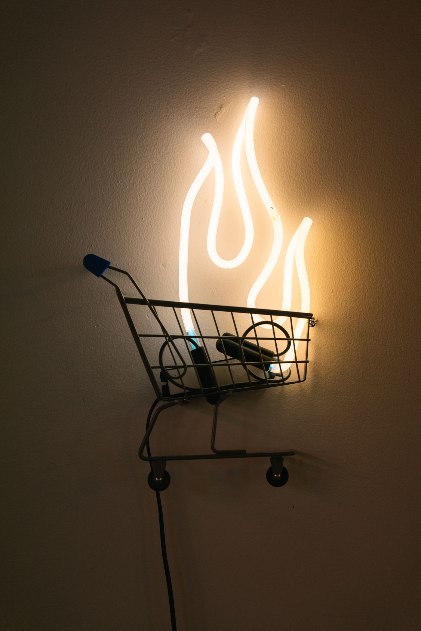 Neon blue flames above a small shopping cart, mounted on a wall; art by Yale Wolf.