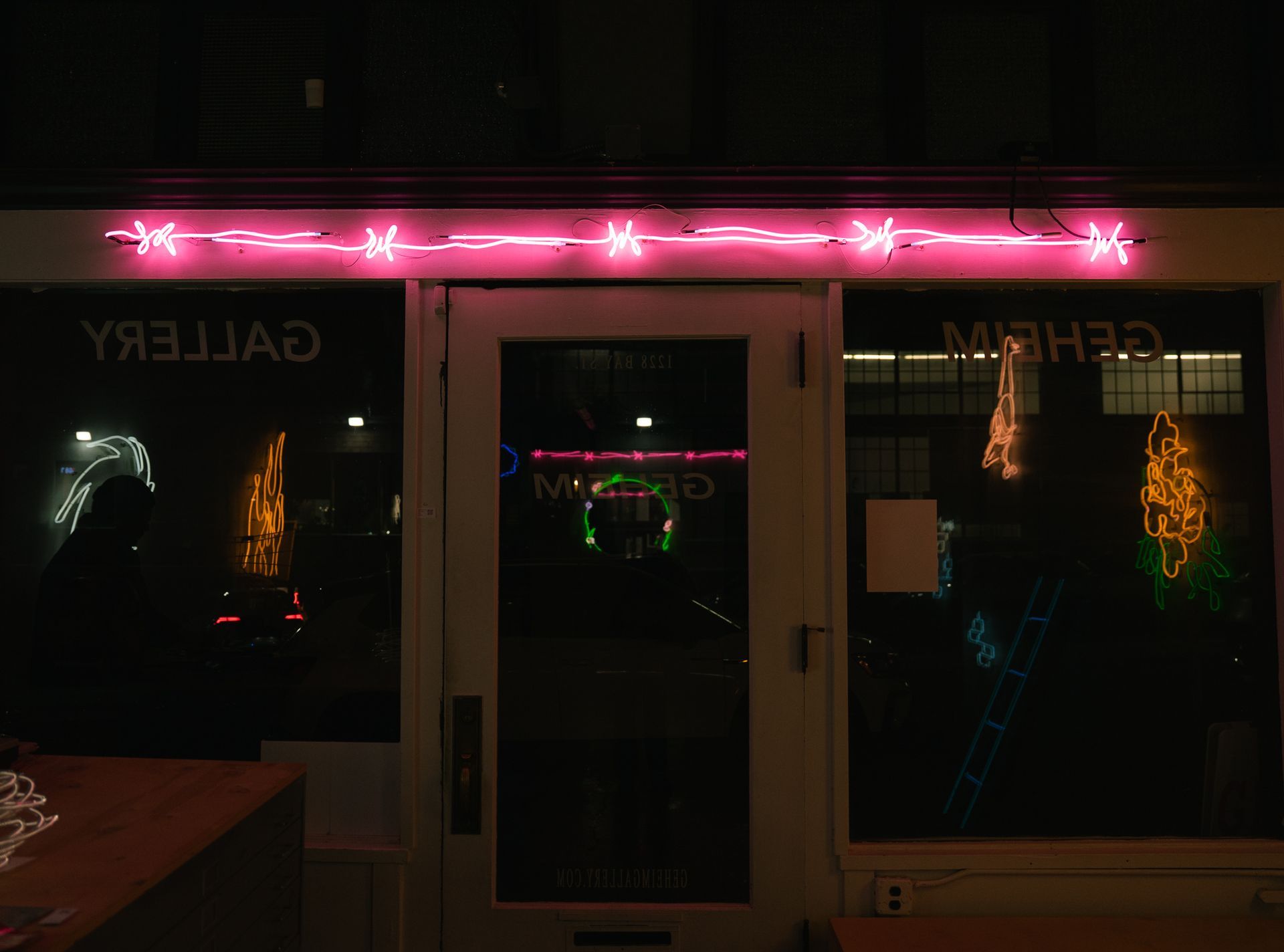 Pink neon lights above a doorway with a reflected logo. A blue light is visible in the background; art by Yale Wolf.