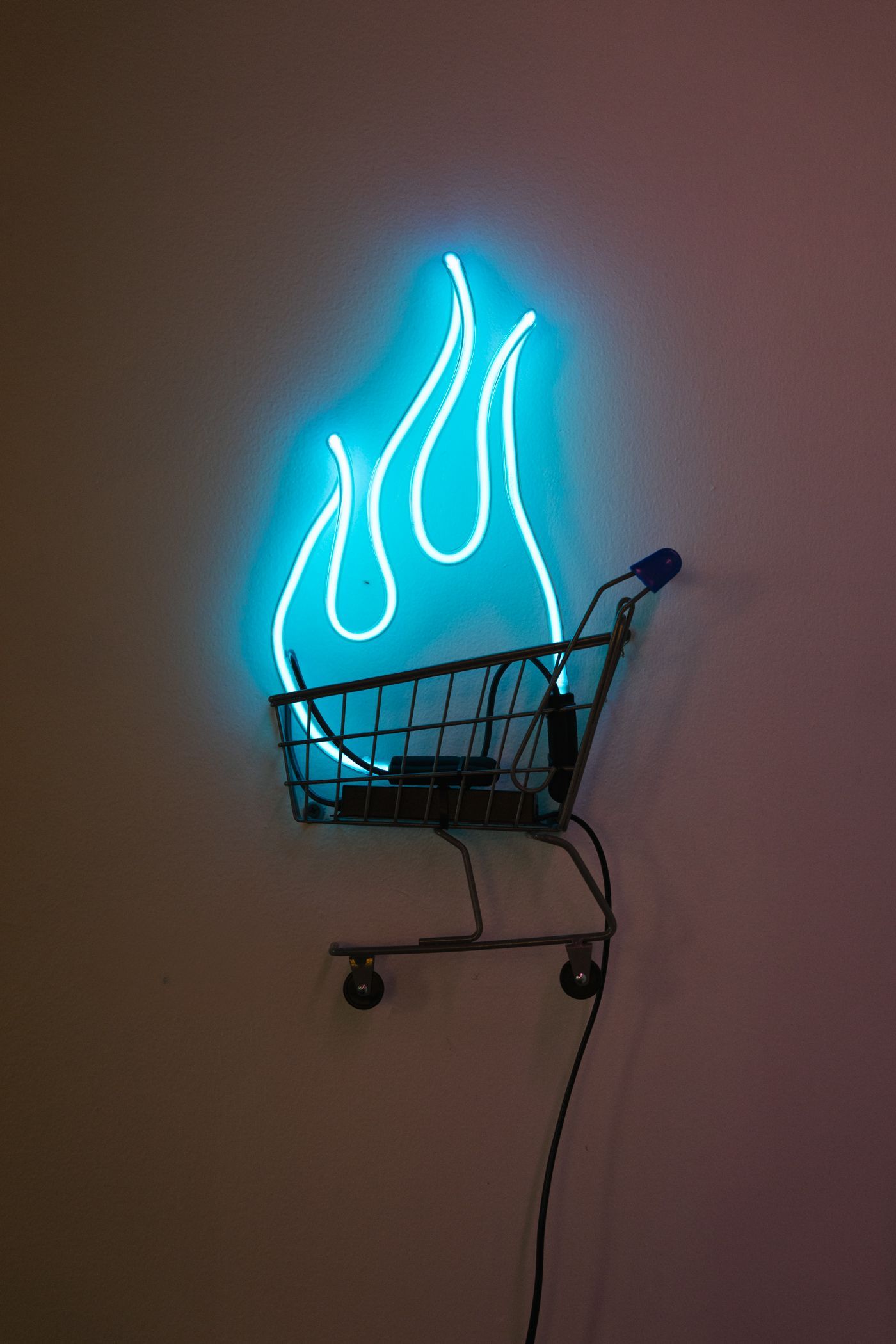 Neon blue flames above a small shopping cart, mounted on a wall; art by Yale Wolf.