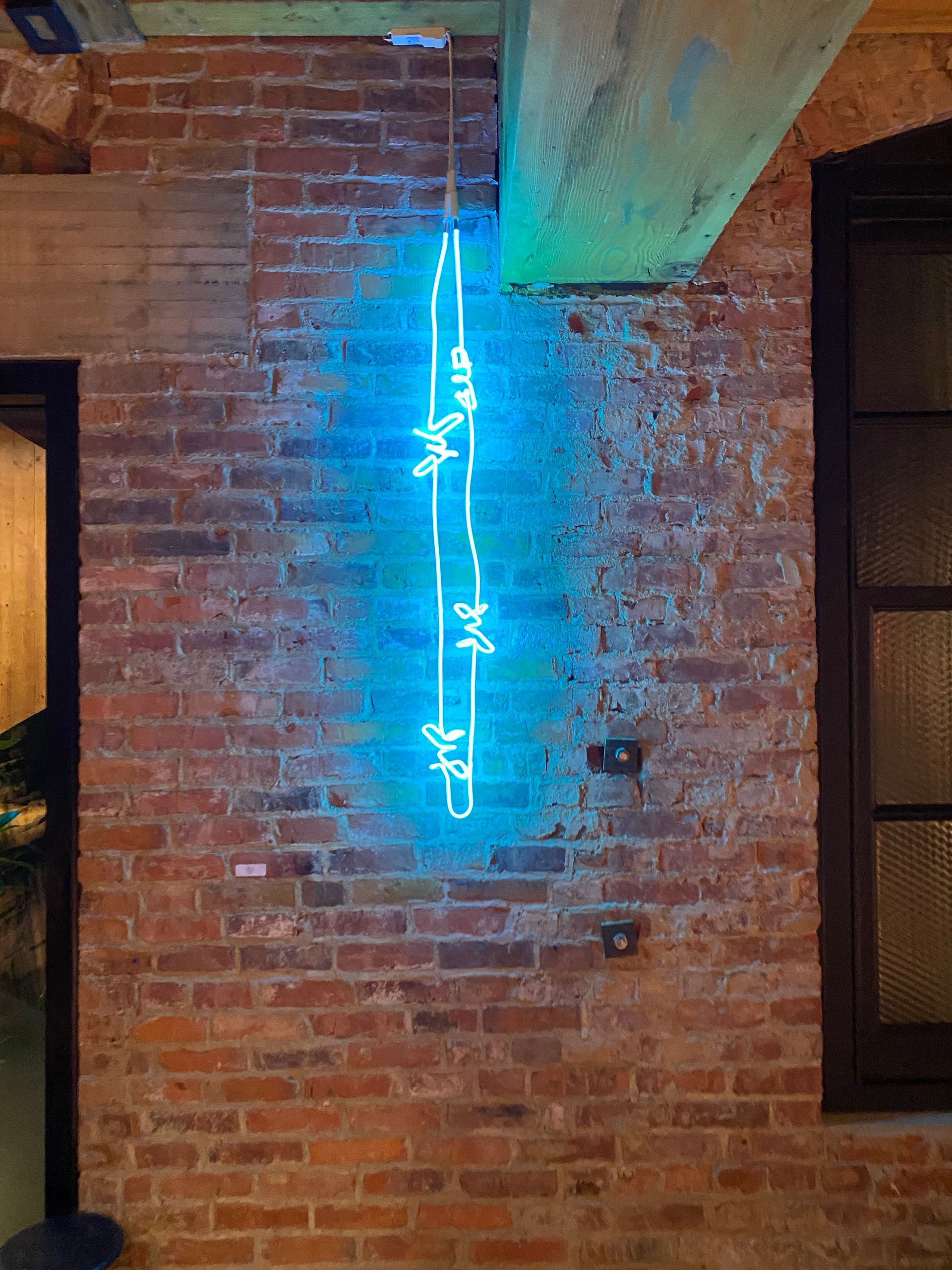 A bright blue neon light shaped like a strand of barbed wire hangs vertically against a red brick wall. Art by Yale Wolf.