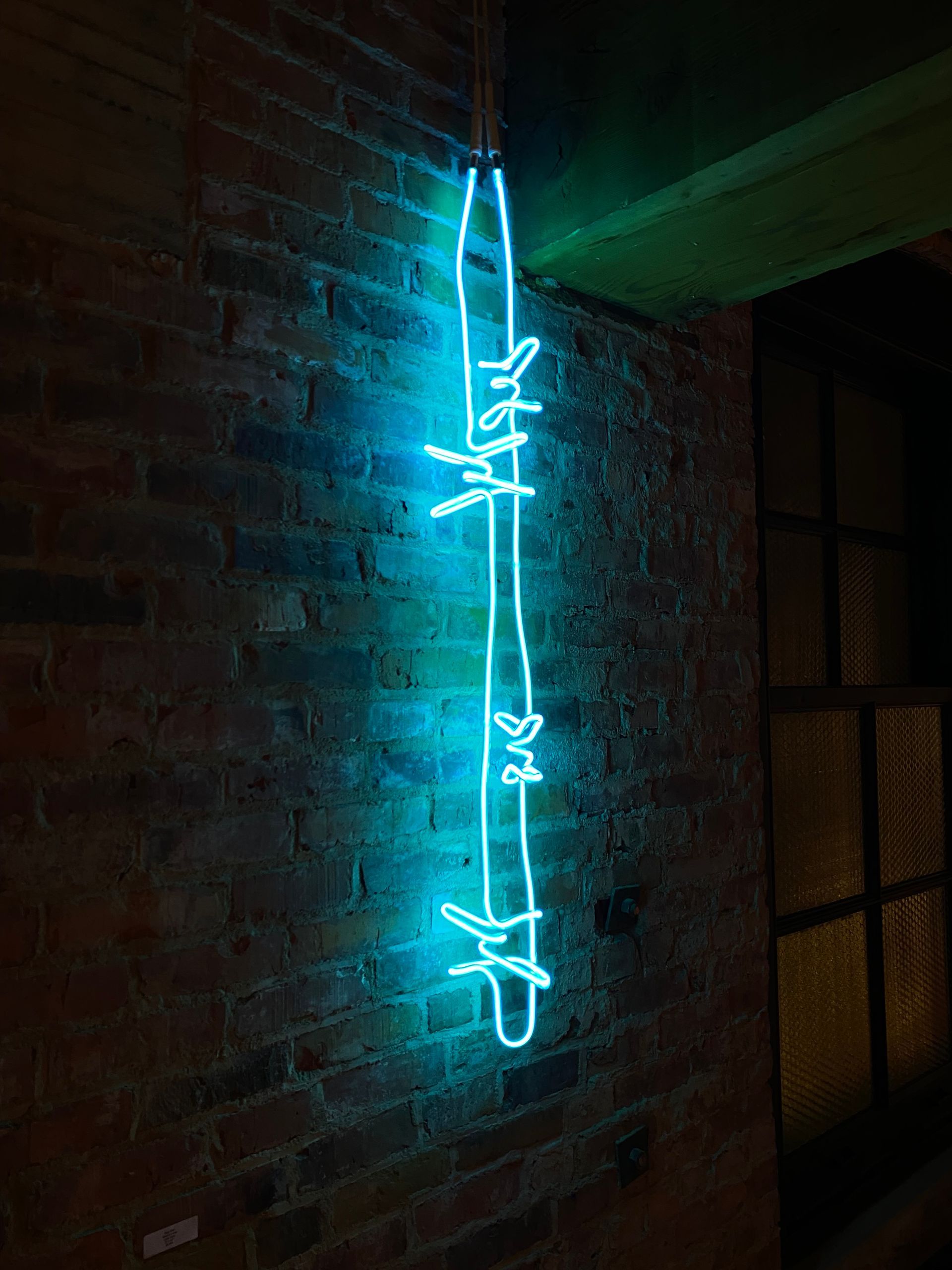 A bright blue neon light shaped like a strand of barbed wire hangs vertically against a red brick wall. Art by Yale Wolf.