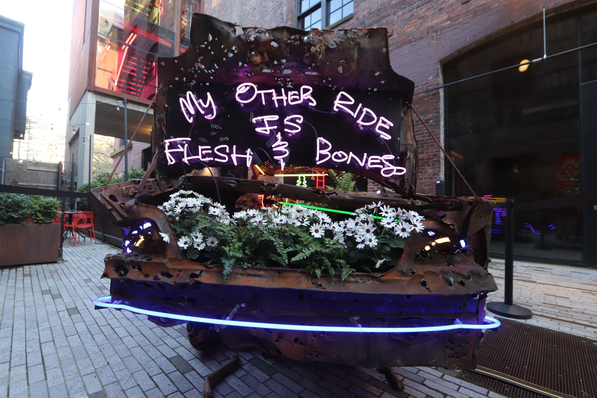 A rusty sculpture with a neon sign that reads "My Other Ride Is Flesh & Bones" over a bed of flowers; by Yale Wolf.