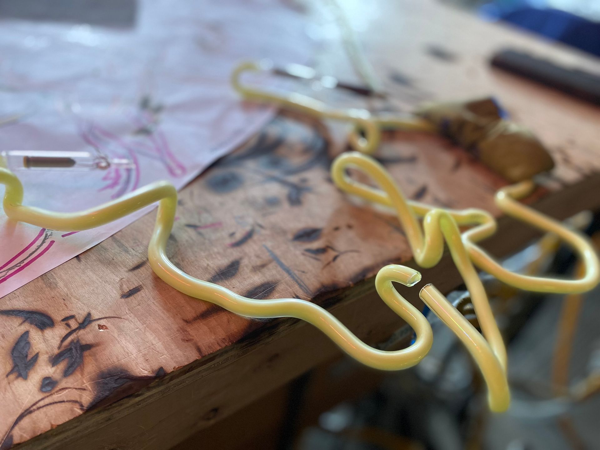A curved length of light yellow neon tubing by Yale Wolf rests on a workbench, positioned above a sketched design on paper.