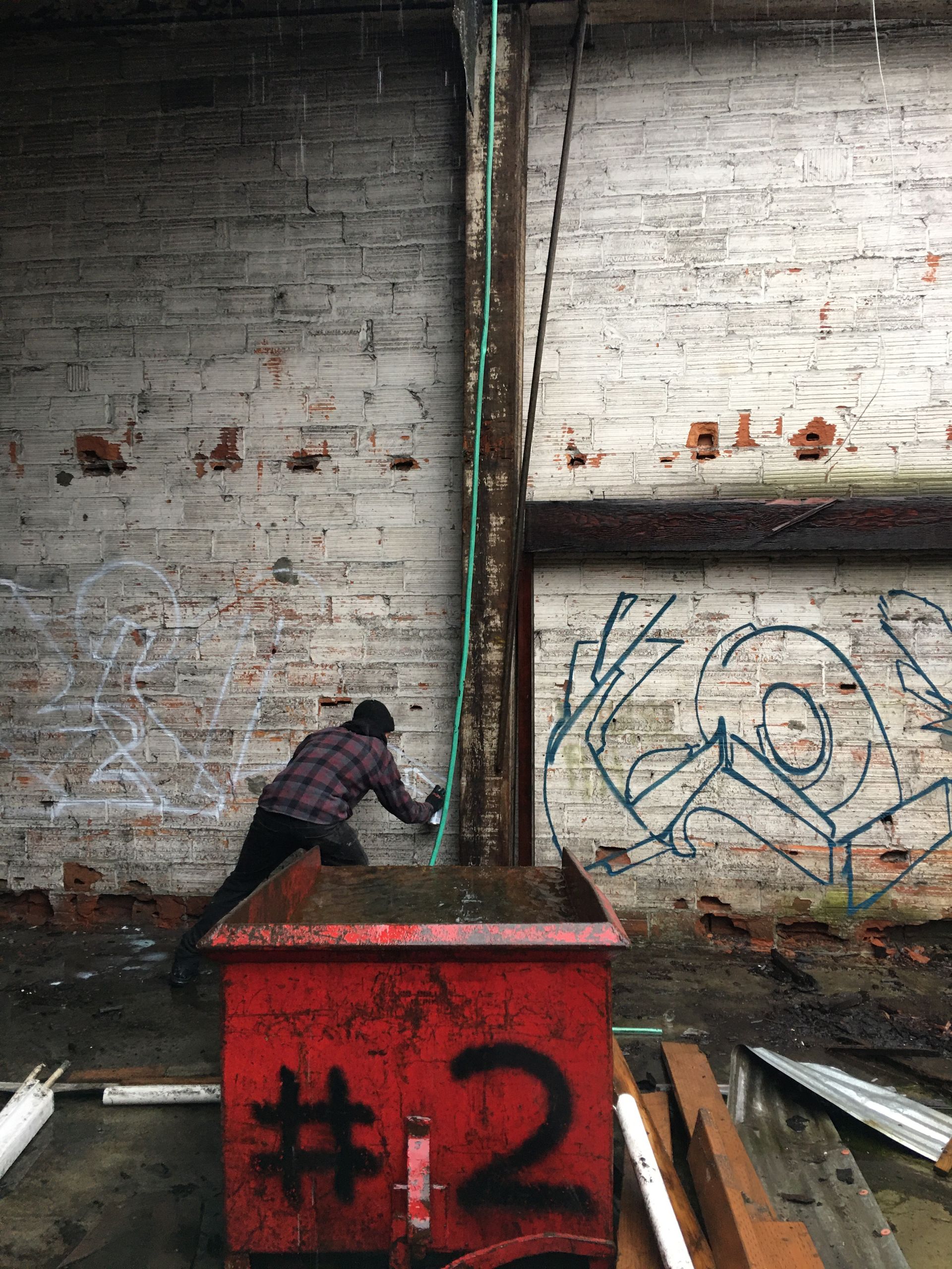 Person spraying something on a metal beam inside an old brick building; red bin in front.