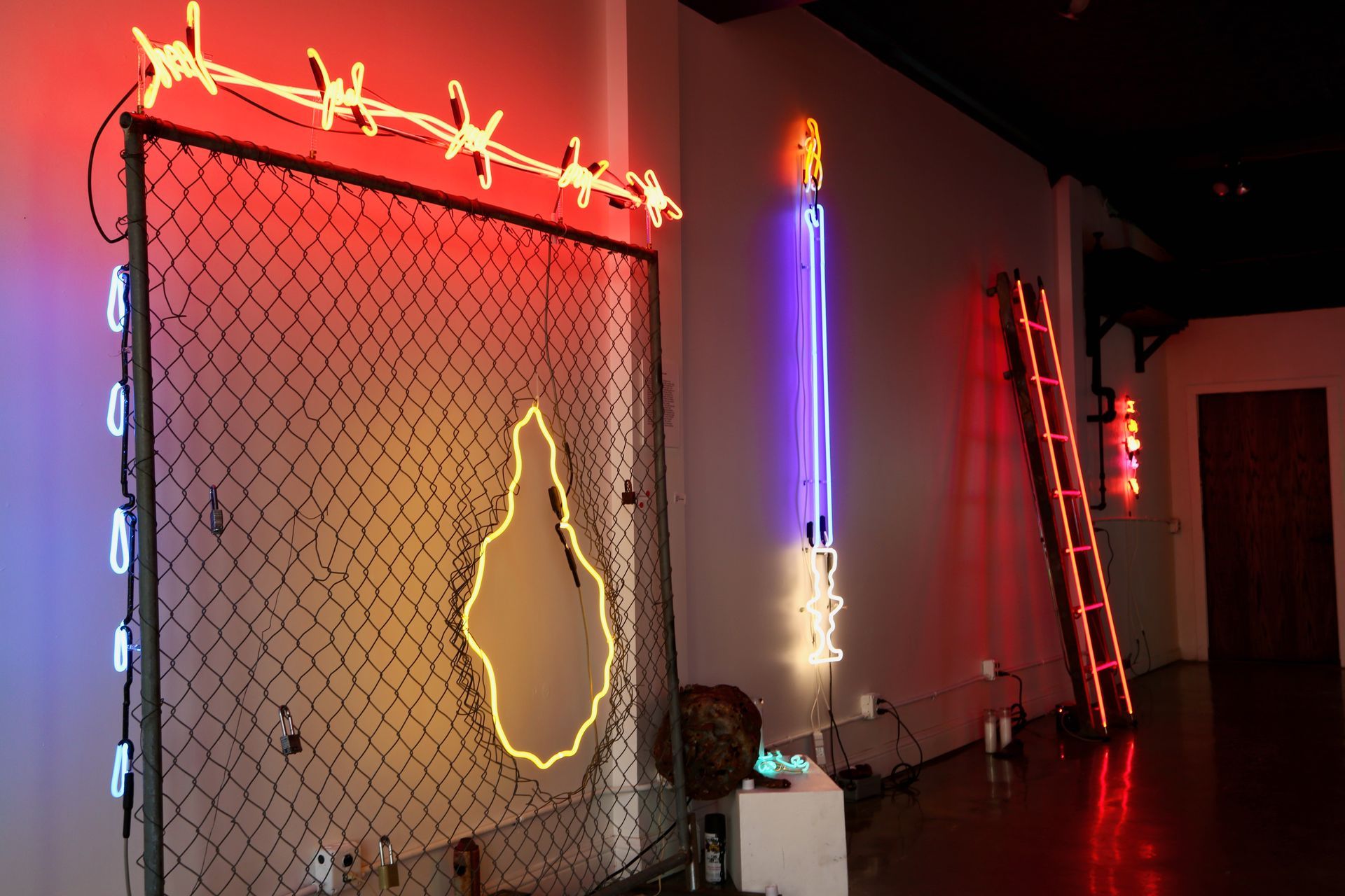 Neon art installation by Yale Wolf: fence with barbed wire, flame, ladder, and vertical tubes in various colors.