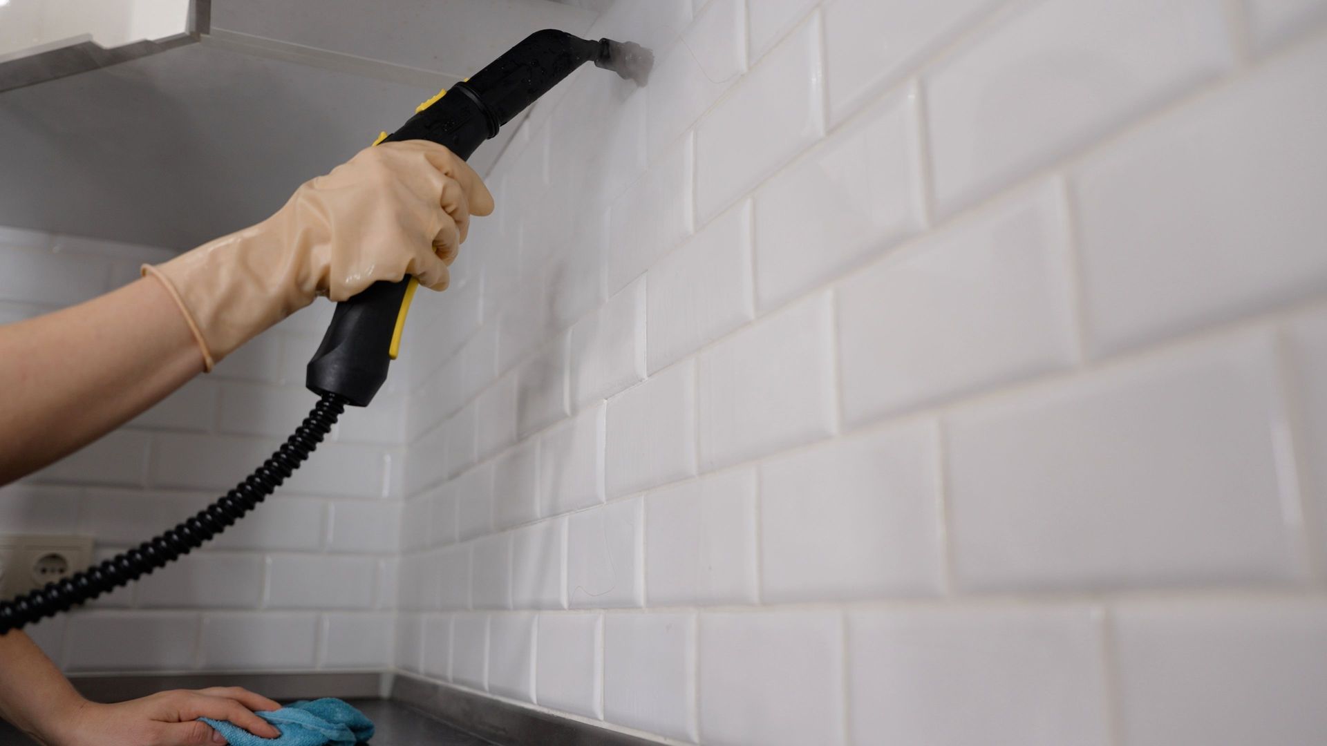 Hand holding a steam cleaner against a white tiled wall in a kitchen