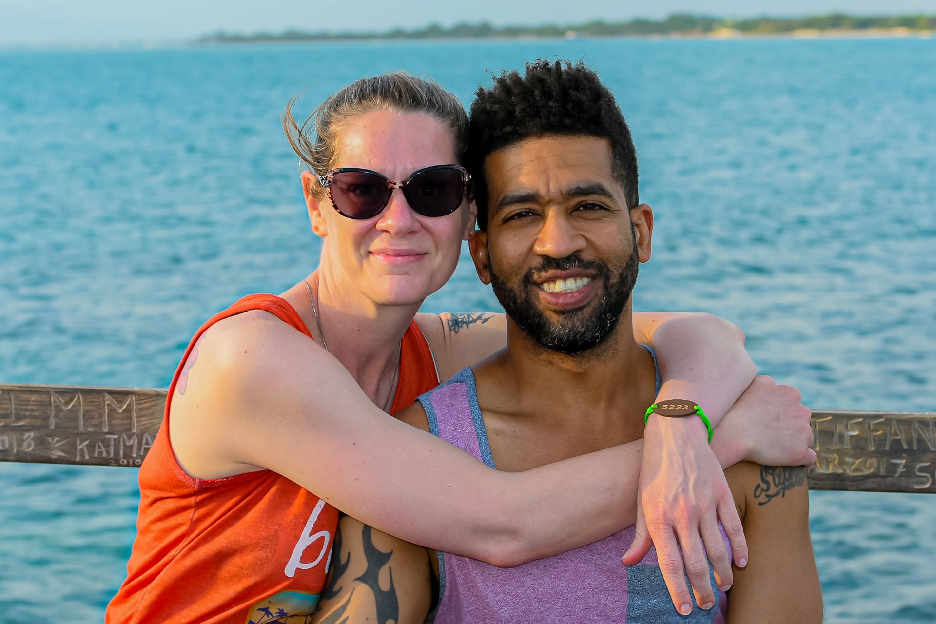 Woman with sunglasses embraces a smiling man by the ocean. Both are looking at the camera.