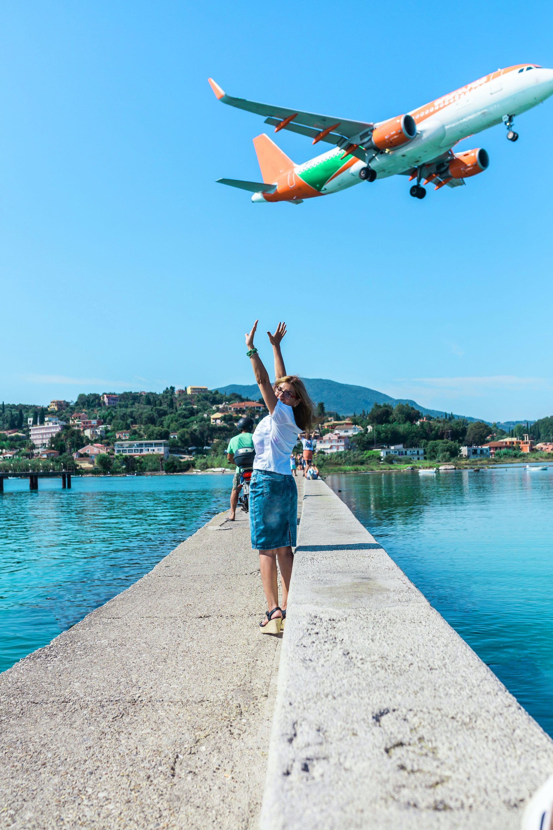 Woman on a pier reaching toward a low-flying orange and white airplane over turquoise water.