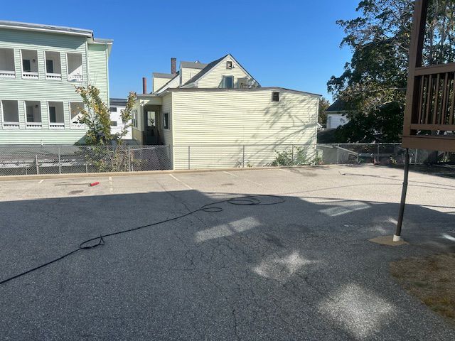 Back yard with a light yellow building, fence, and paved surface.