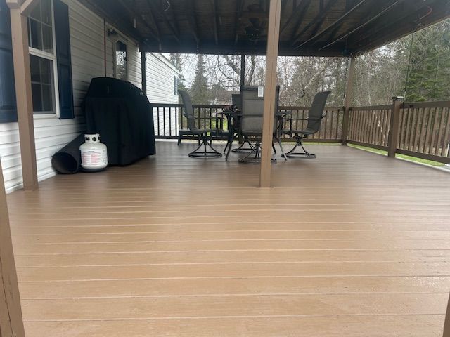 Covered backyard deck with tan flooring, railing, patio table and chairs, and a covered grill.