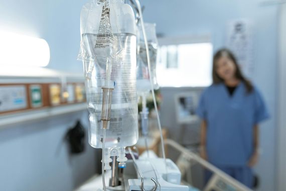 An intravenous drip bag hanging in a hospital room with a medical professional out of focus in the background.