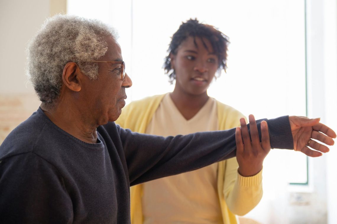 A healthcare provider in a yellow cardigan assists an individual with physical therapy exercises by holding their wrist.