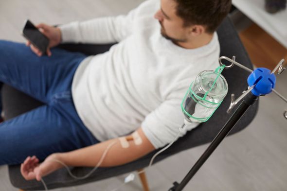 A person sits in a chair using a phone while receiving an intravenous infusion from a hanging bag.