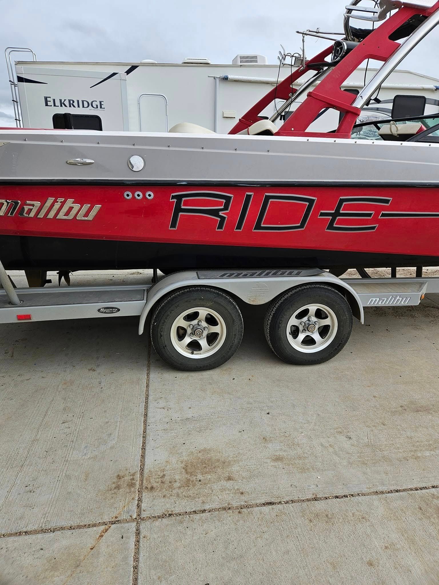 A side view of a red and silver Malibu boat on a dual-axle trailer, parked on a concrete surface.