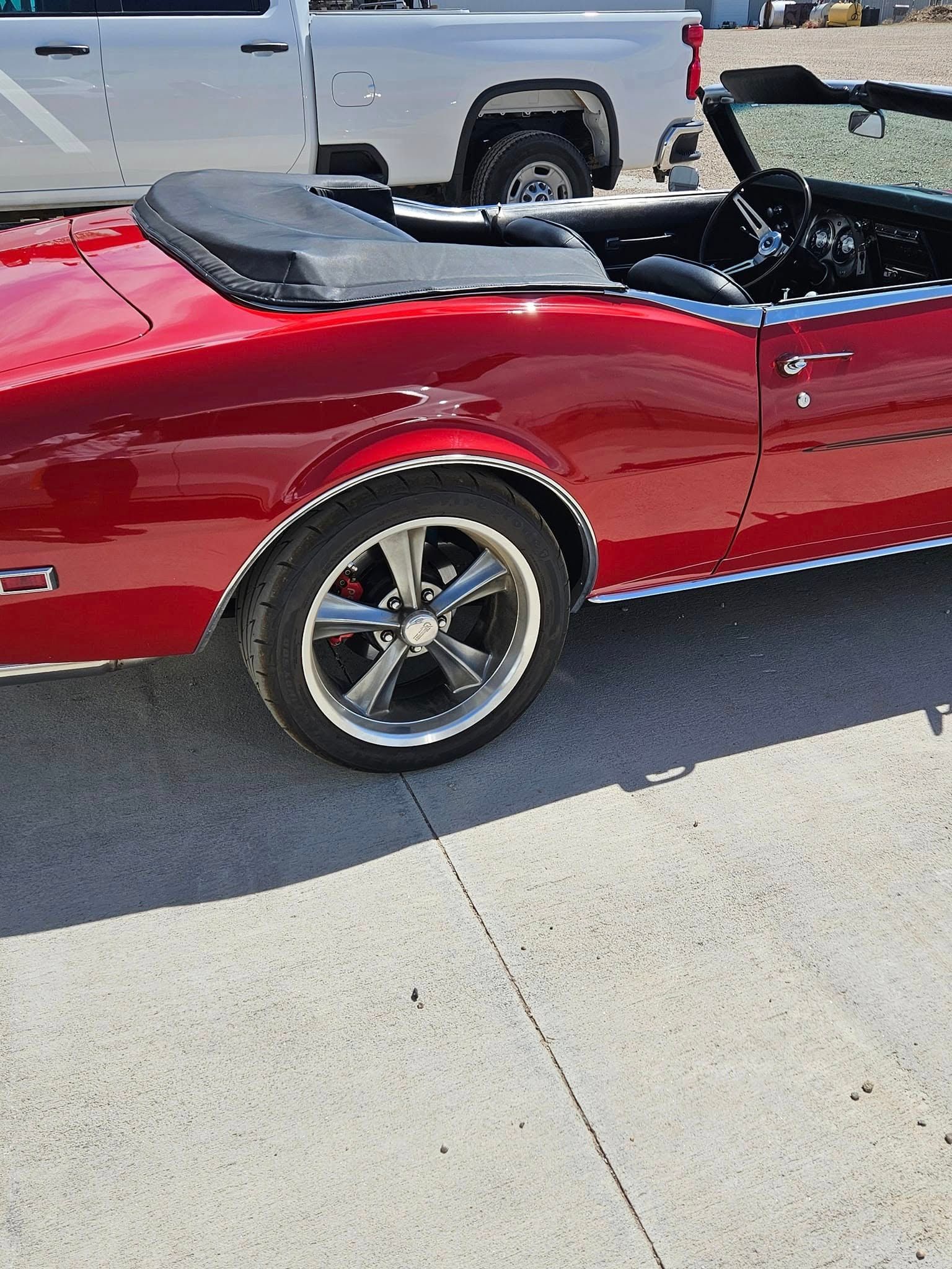 A bright red vintage convertible car parked on a paved lot with a silver pickup truck in the background.