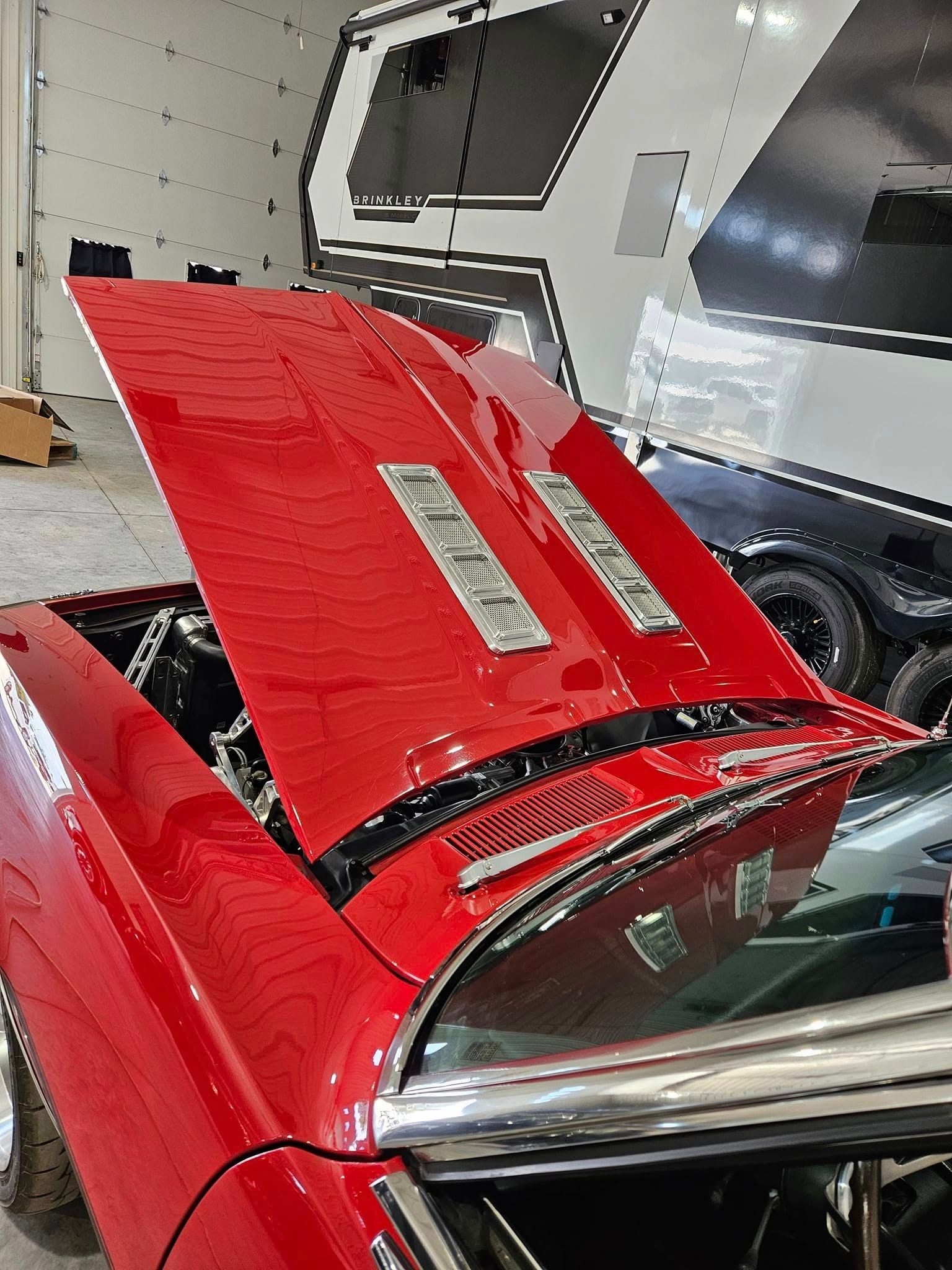 A red muscle car with its hood open, showing chrome vents, parked inside a garage next to a large trailer.