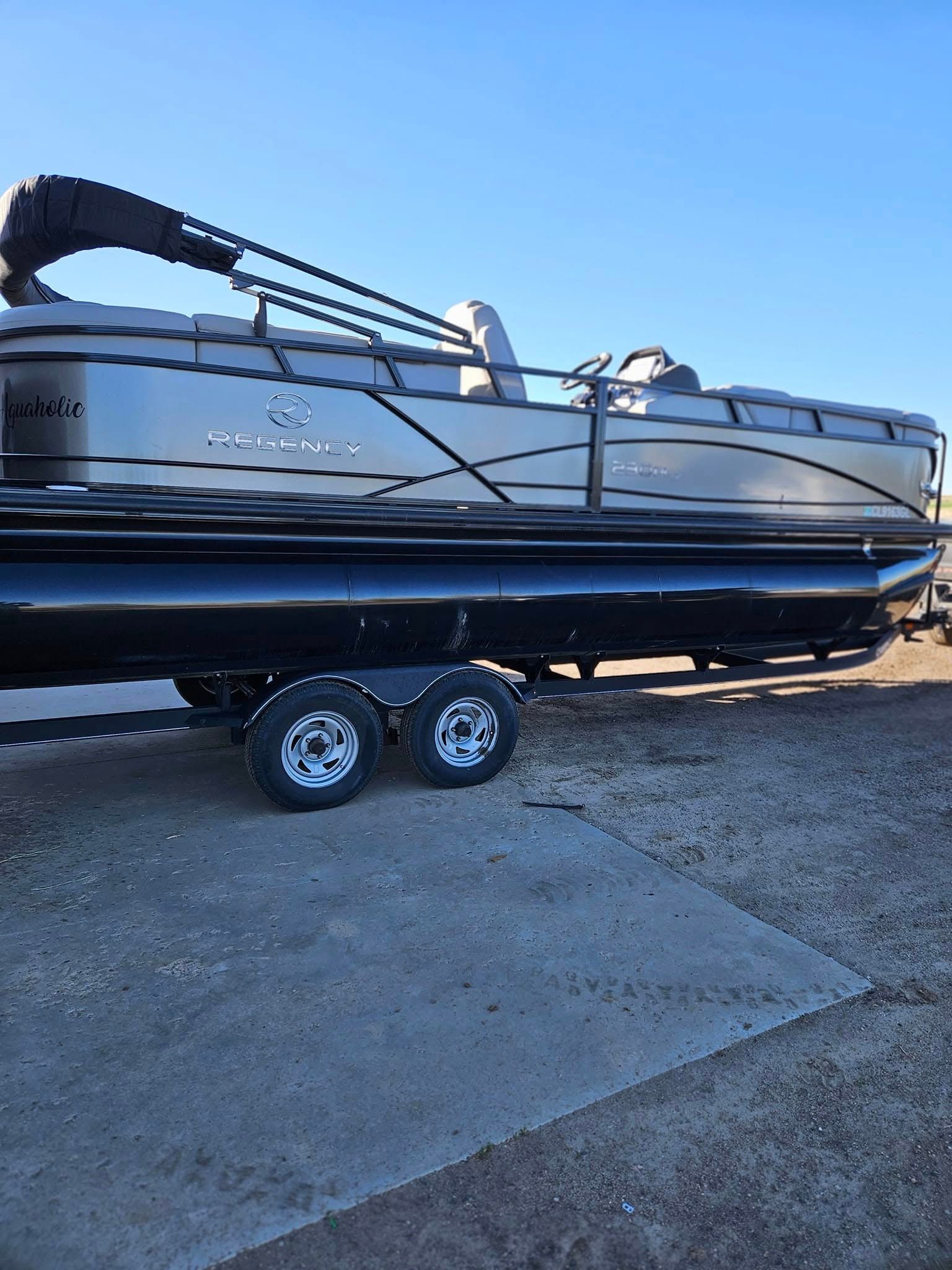 A silver and black pontoon boat on a double-axle trailer, parked on a gravel surface under a clear blue sky.