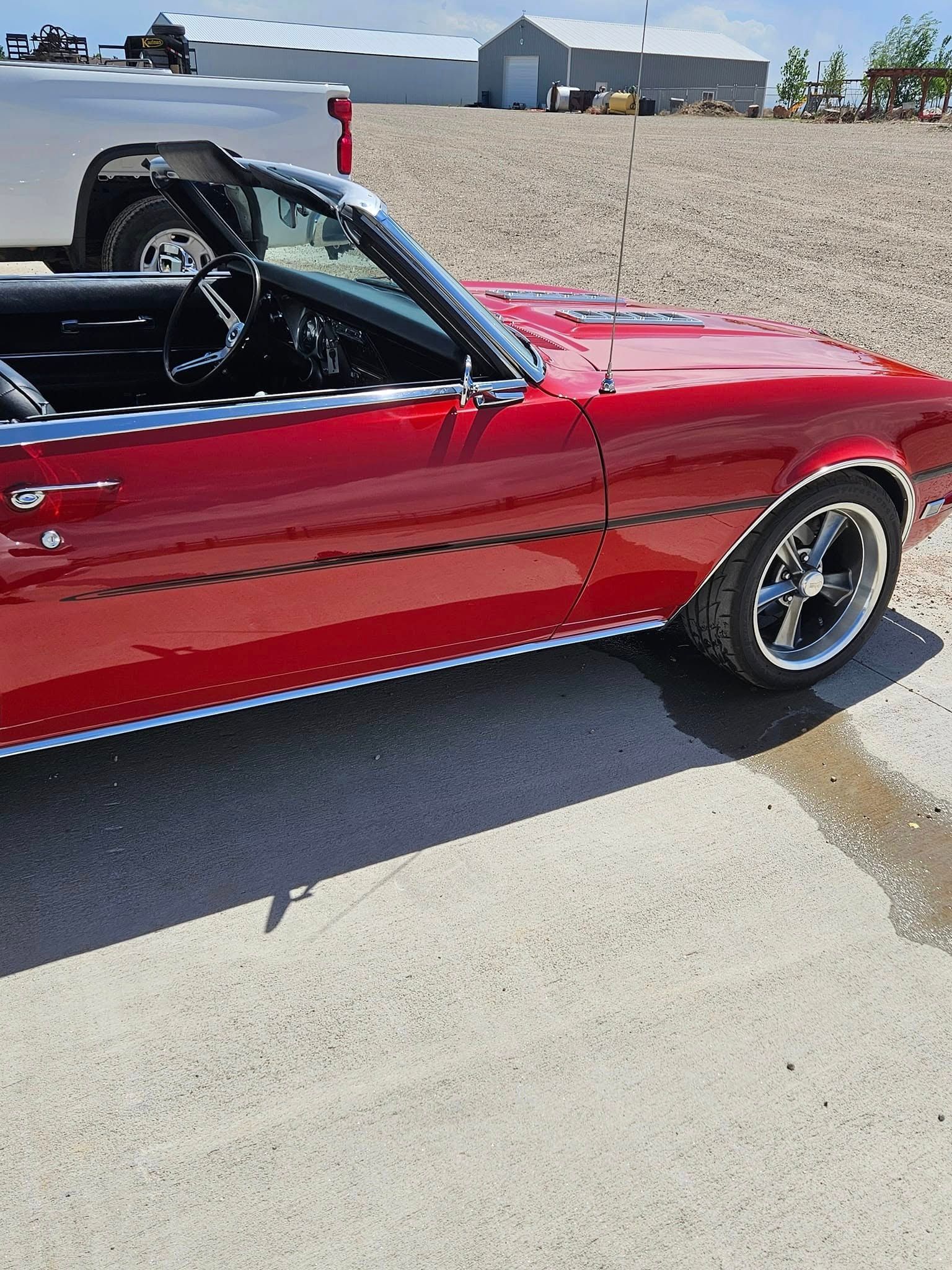 A shiny, bright red classic convertible parked outdoors on a gravel lot next to a white truck.