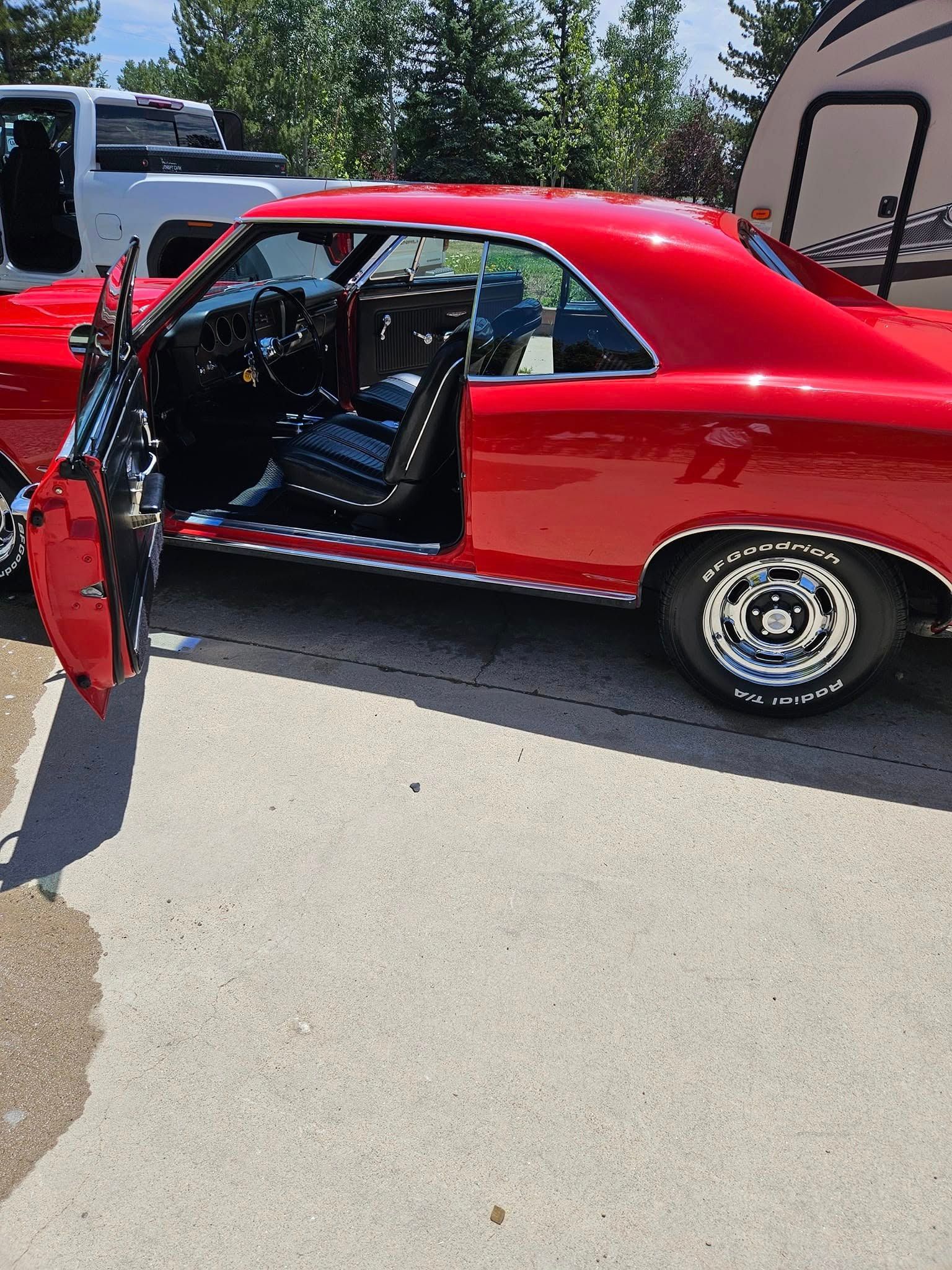 A bright red classic muscle car with the driver’s door open, parked on a paved lot with a white truck and camper nearby.