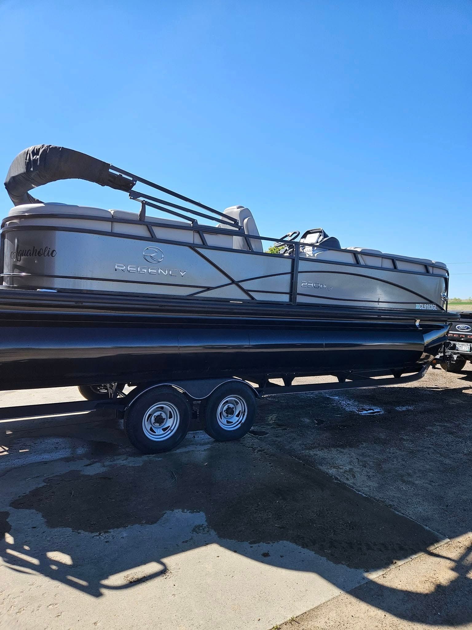 A black pontoon boat with a folded canopy sits on a tandem-axle trailer against a clear blue sky.