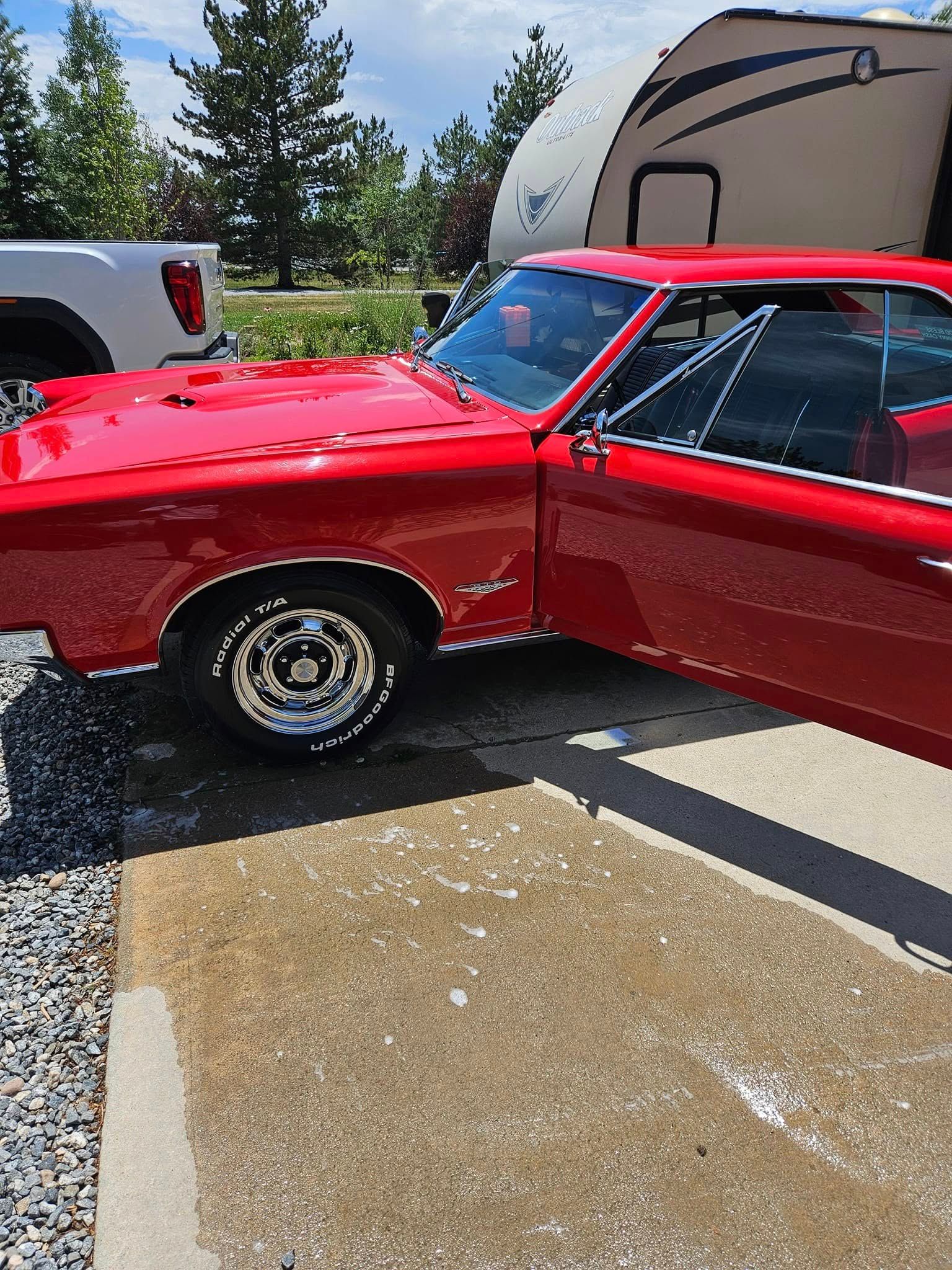 A bright red vintage muscle car with an open door parked on a concrete driveway next to a white pickup and an RV.