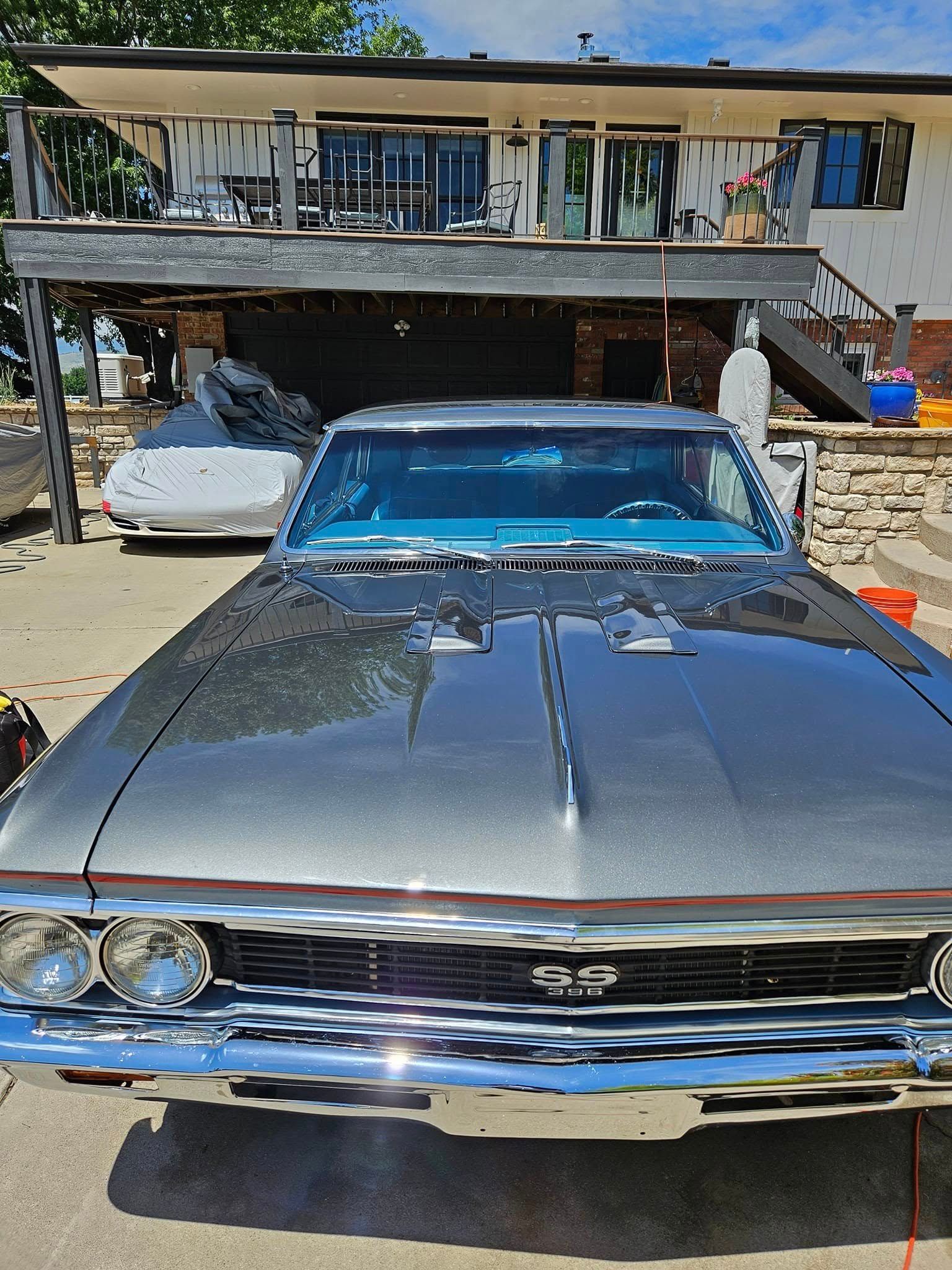A dark metallic classic Chevrolet SS muscle car parked on a concrete driveway in front of a house with a balcony.