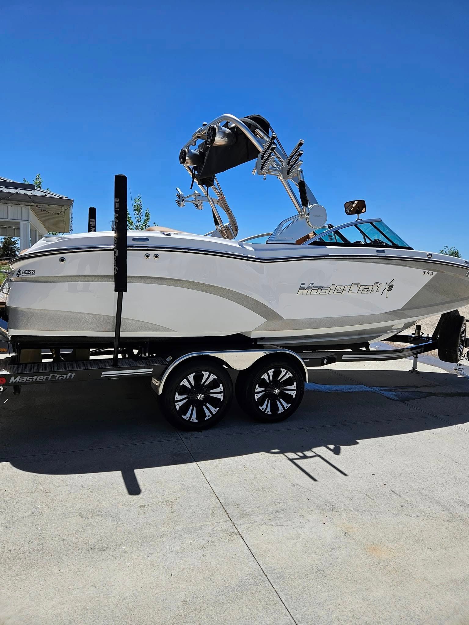 A white motorboat with a black wakeboard tower on a tandem-axle trailer, parked on a concrete surface under a blue sky.