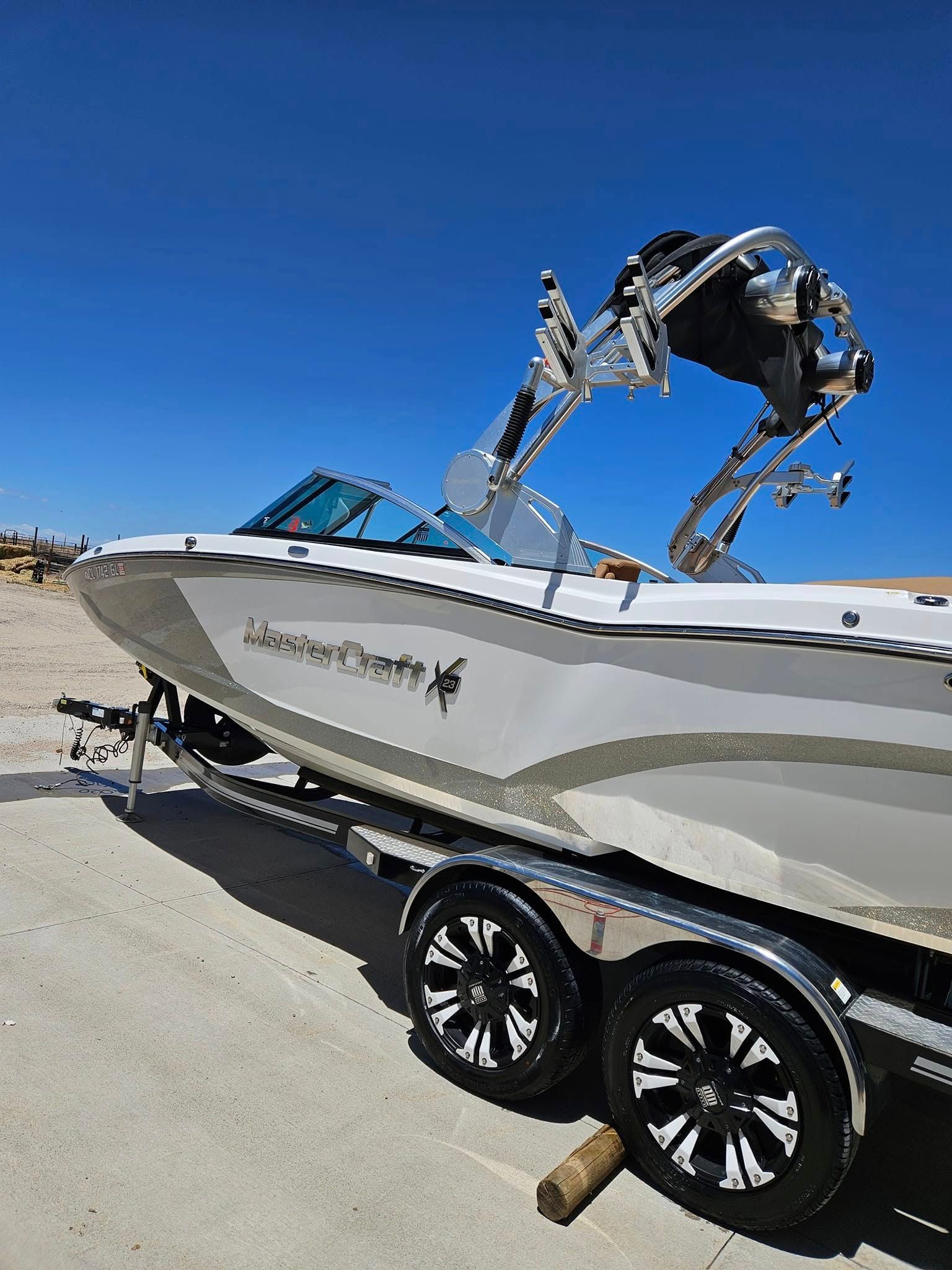 A silver and white boat with a wakeboard tower mounted on a dual-axle trailer, parked on a paved surface under a blue sky.