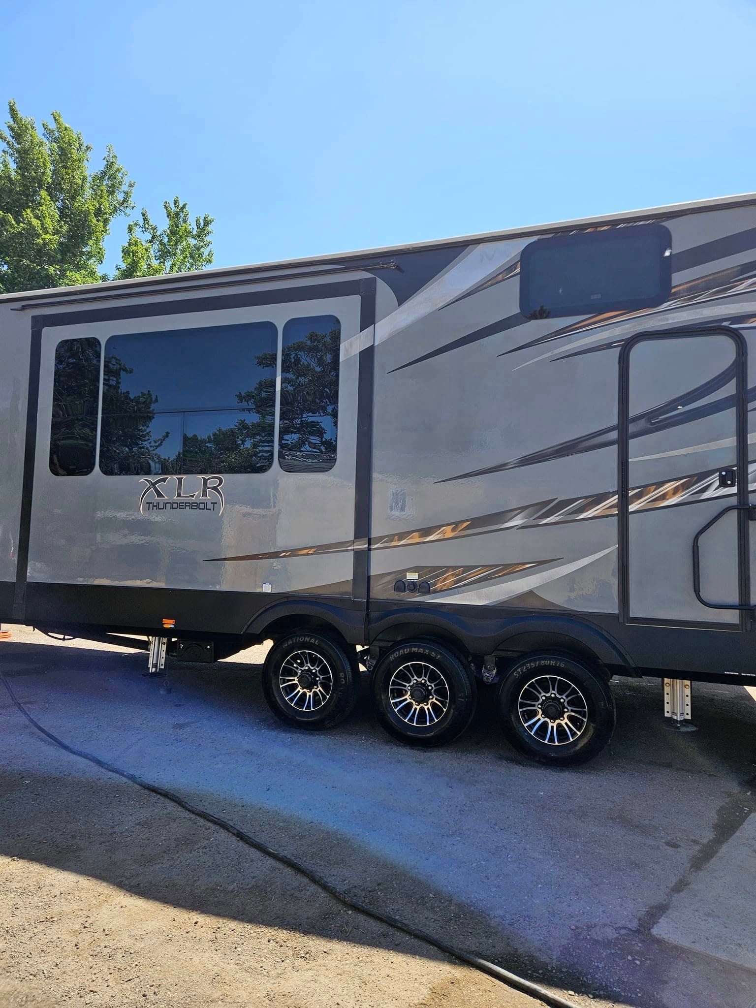 A side view of a modern grey recreational vehicle parked outdoors on a sunny day with three wheels visible.