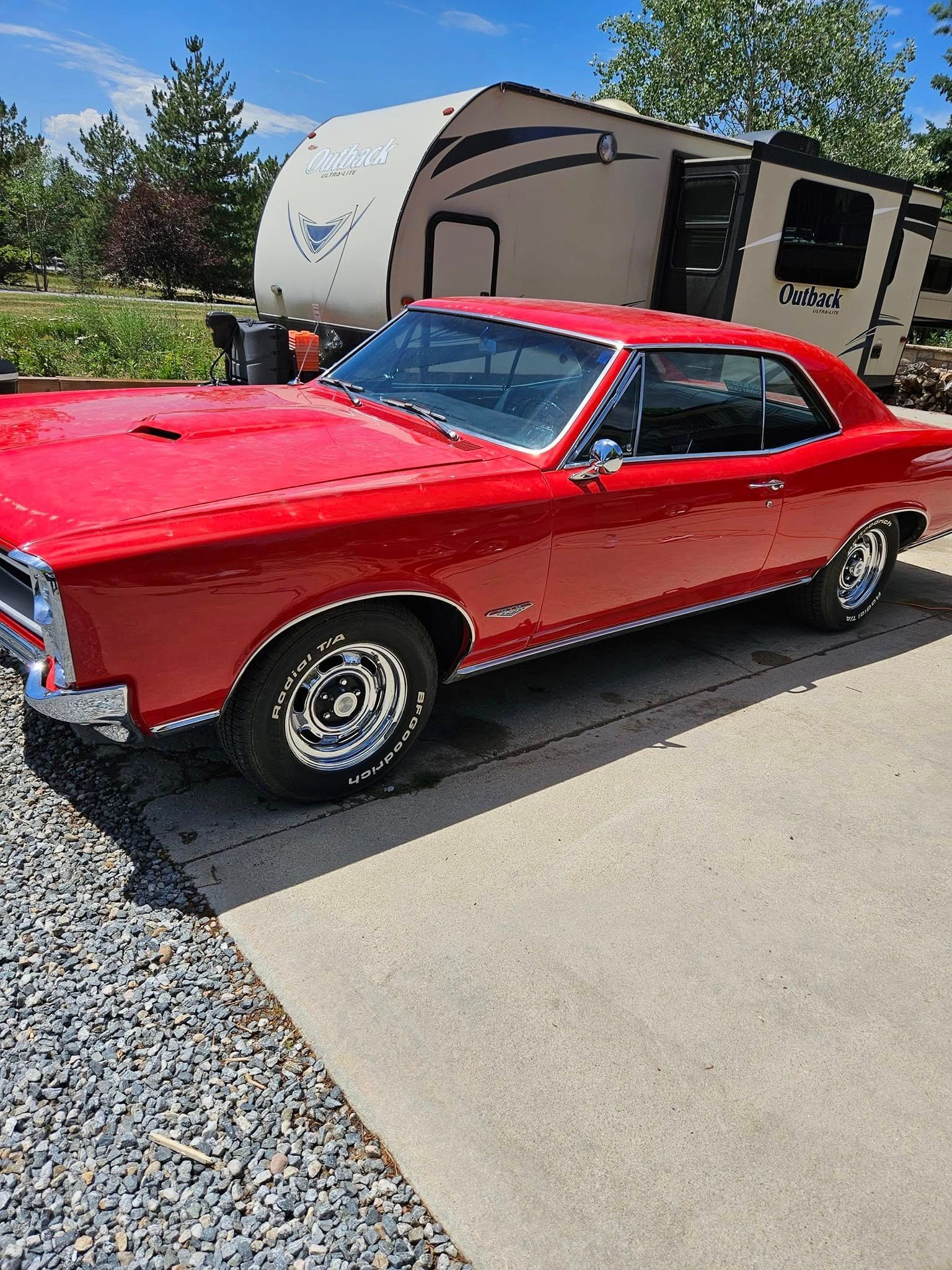 A bright red classic muscle car parked on a concrete driveway in front of a camper trailer.