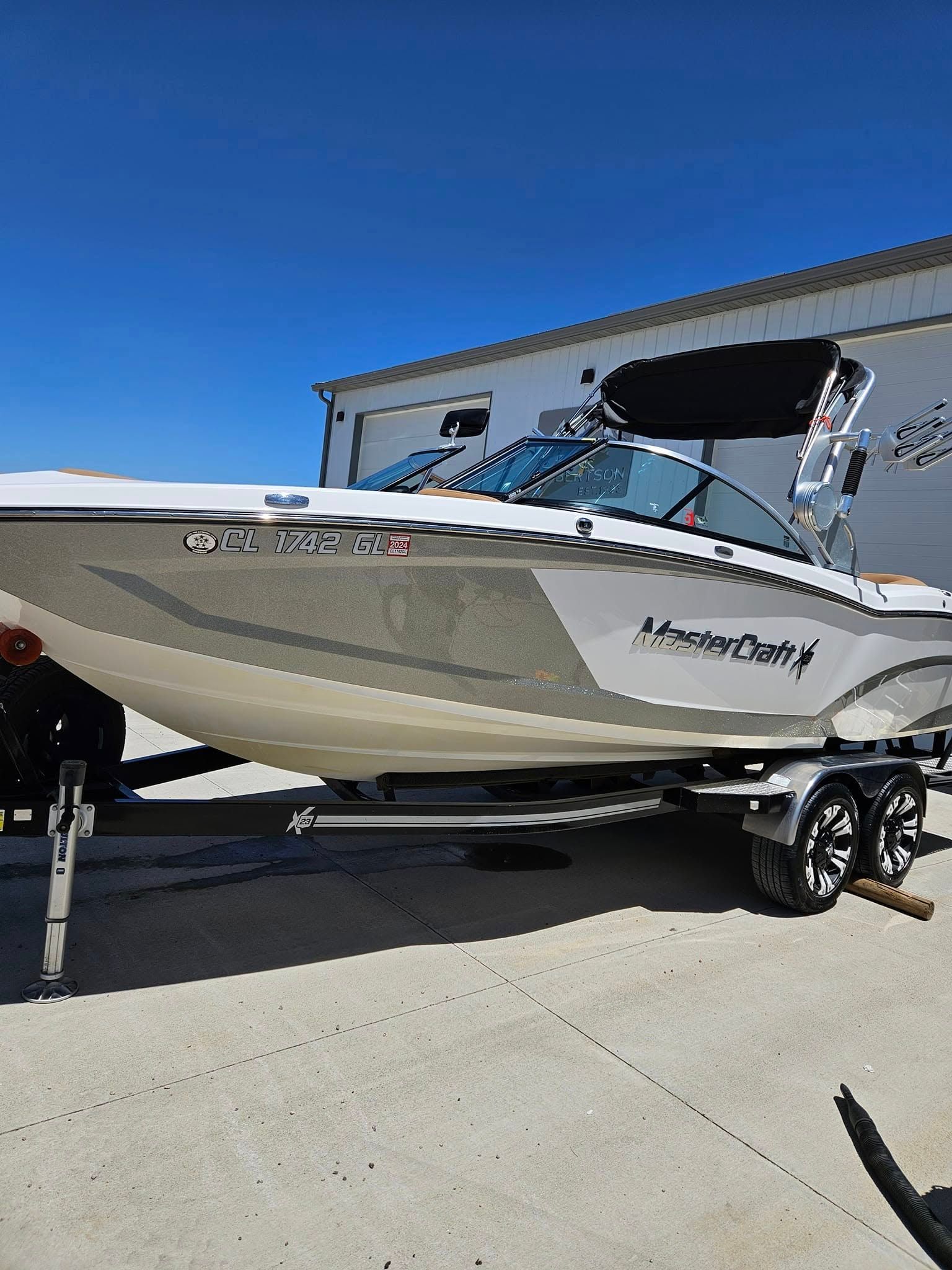 A white and grey MasterCraft powerboat sits on a boat trailer in a concrete lot against a clear blue sky.