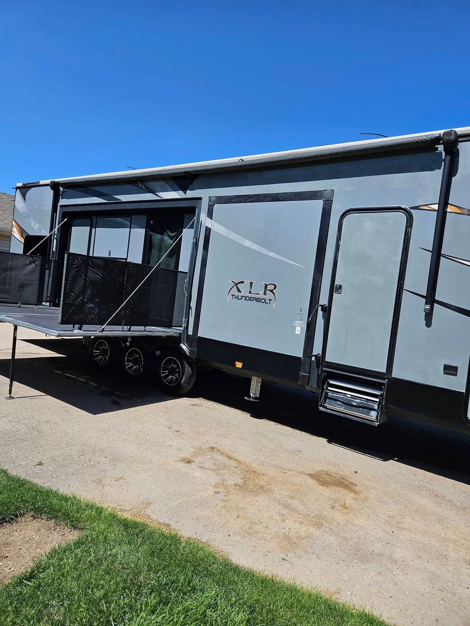 A grey Forest River XLE toy hauler RV parked on gravel with its rear patio ramp lowered and screen walls deployed.