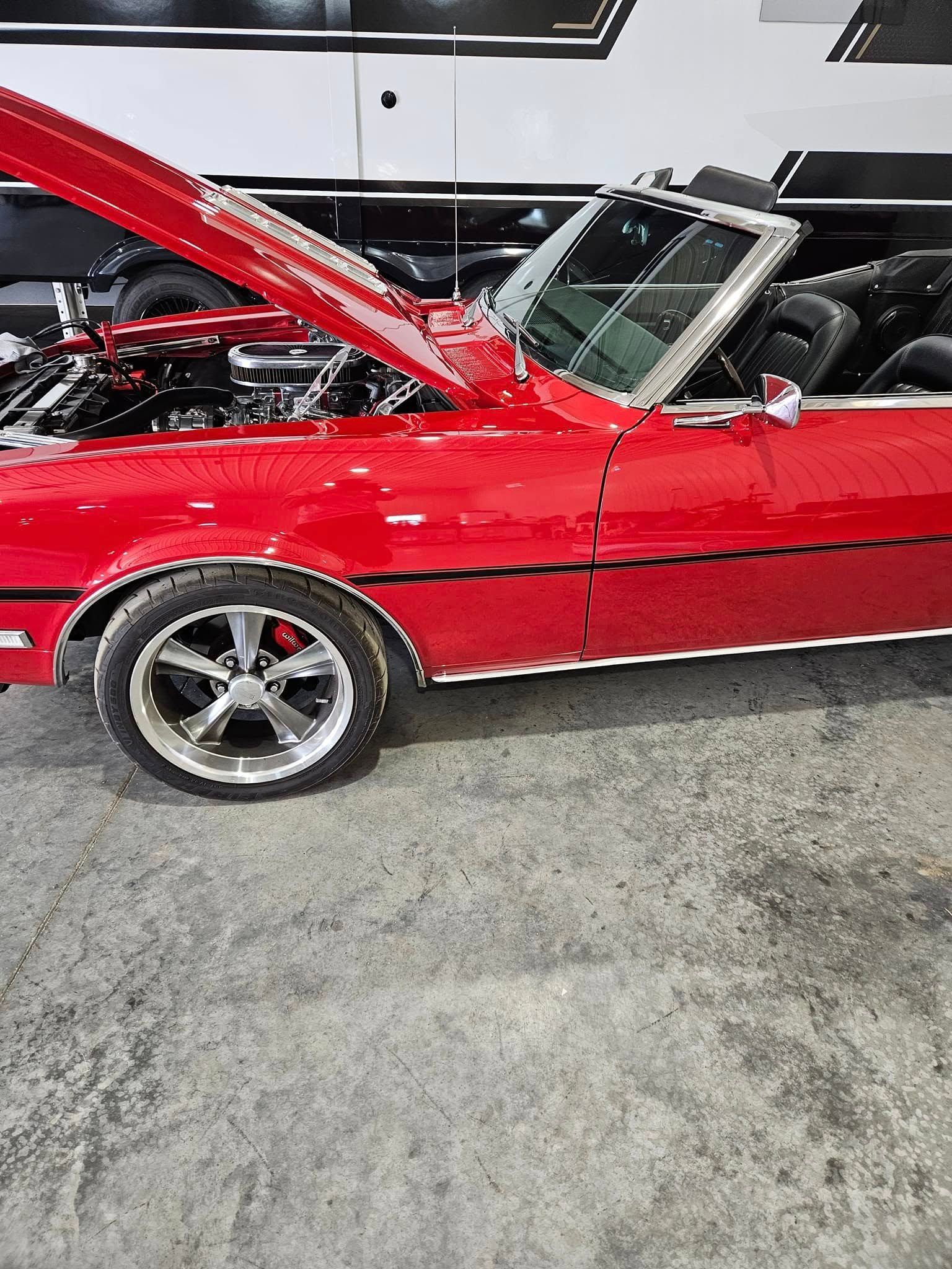 A bright red classic convertible car with its hood open, parked on a concrete floor next to a trailer.