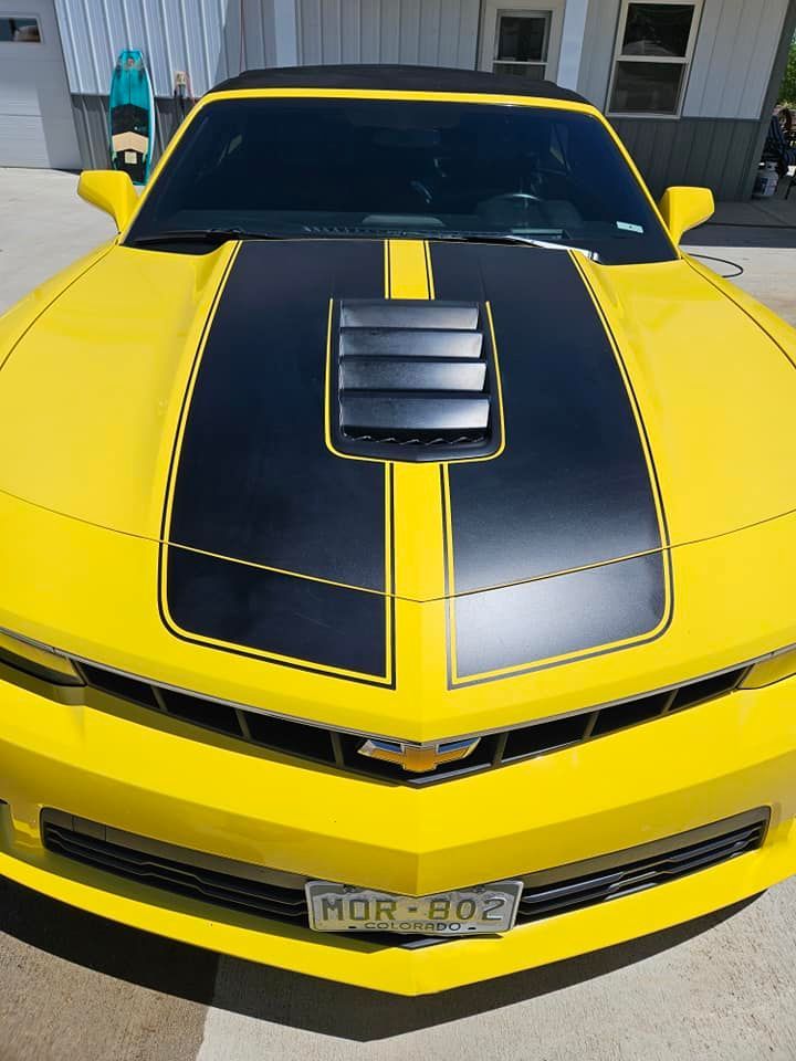A bright yellow Chevrolet Camaro with a matte black hood stripe and vent, parked on concrete in front of a building.