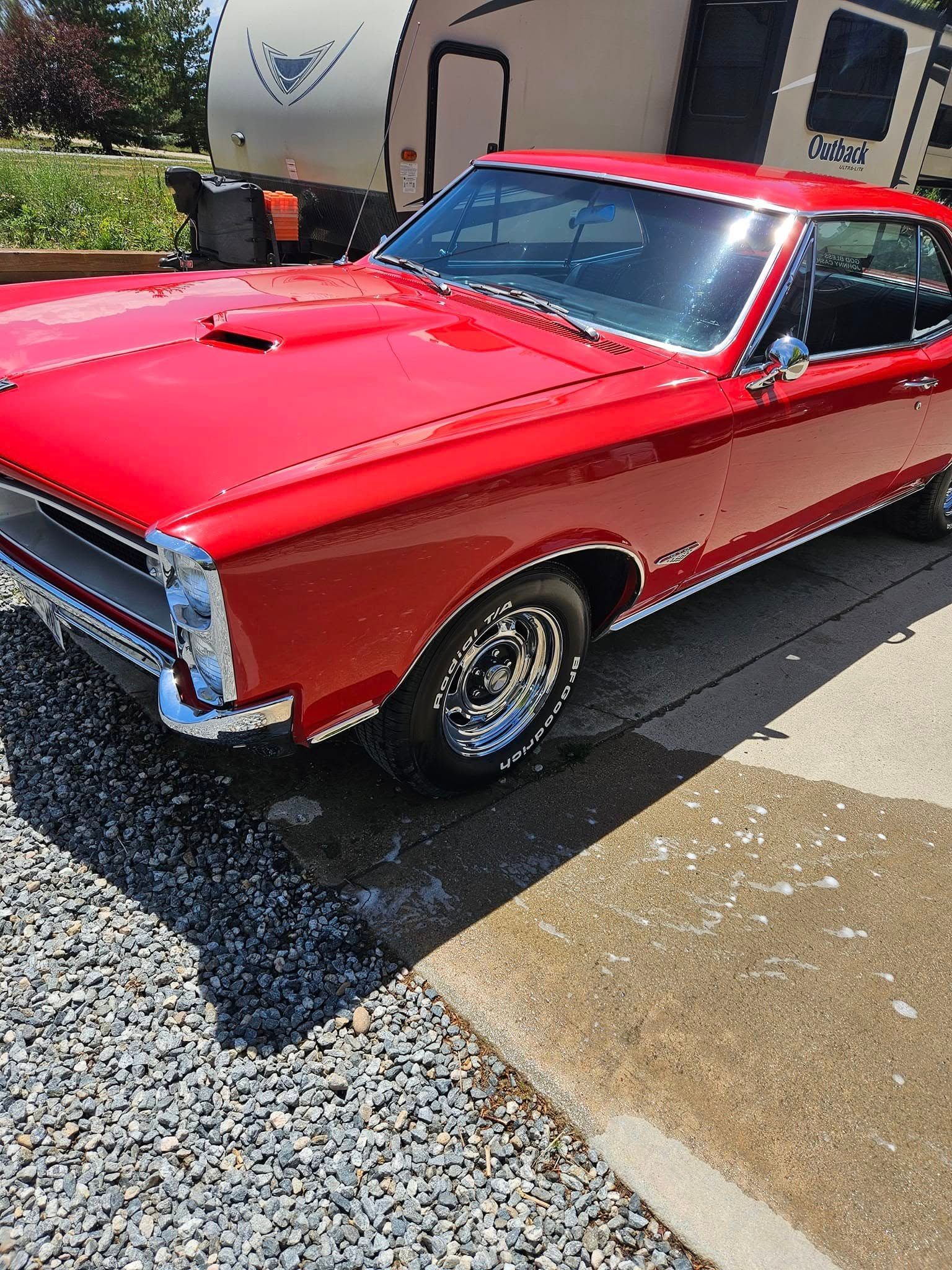 A bright red classic Pontiac GTO muscle car parked outdoors on a sunny day next to a camper trailer.