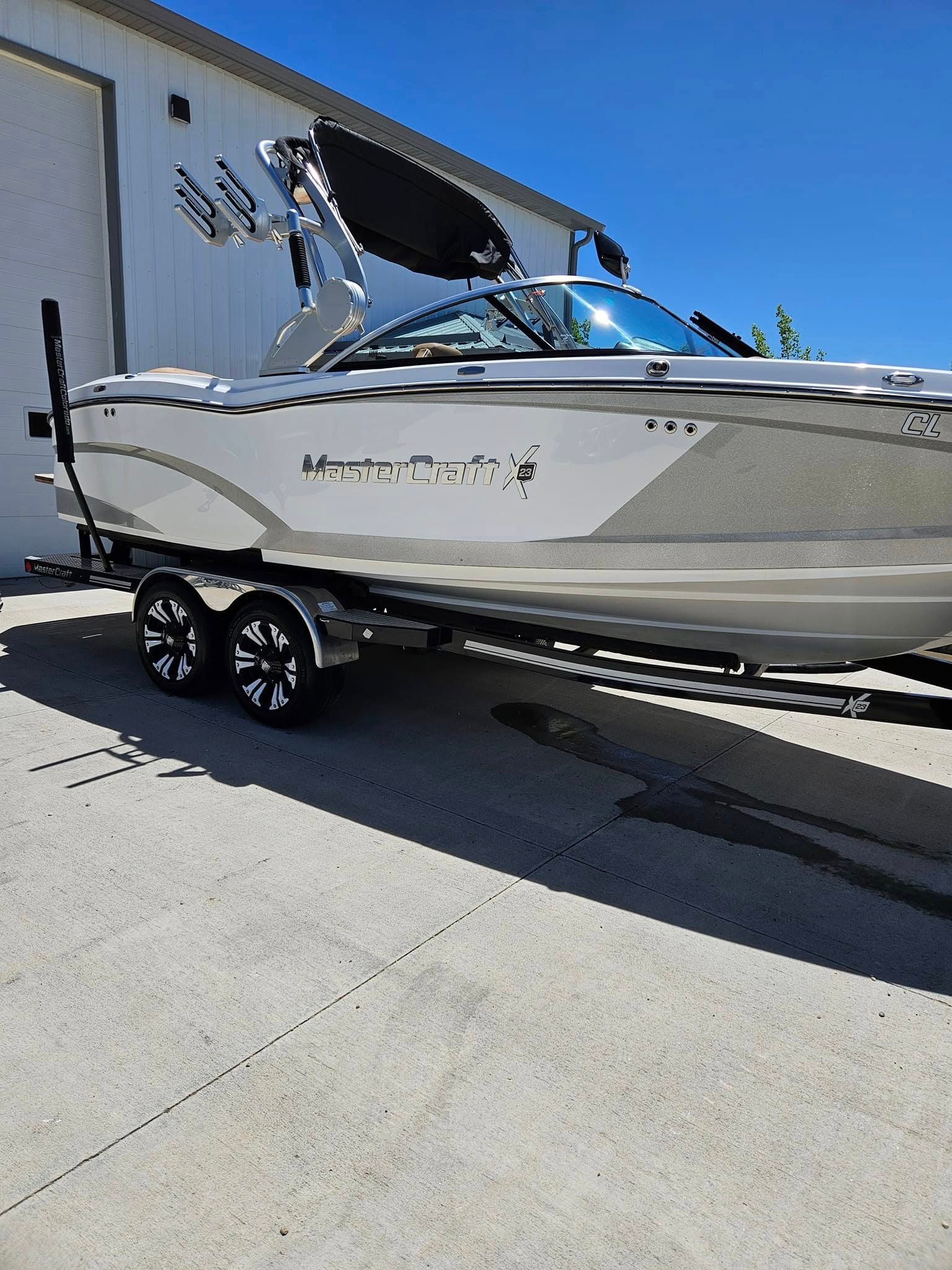 A white and gray MasterCraft motorboat on a tandem-axle trailer, parked on a concrete surface in front of a white building.