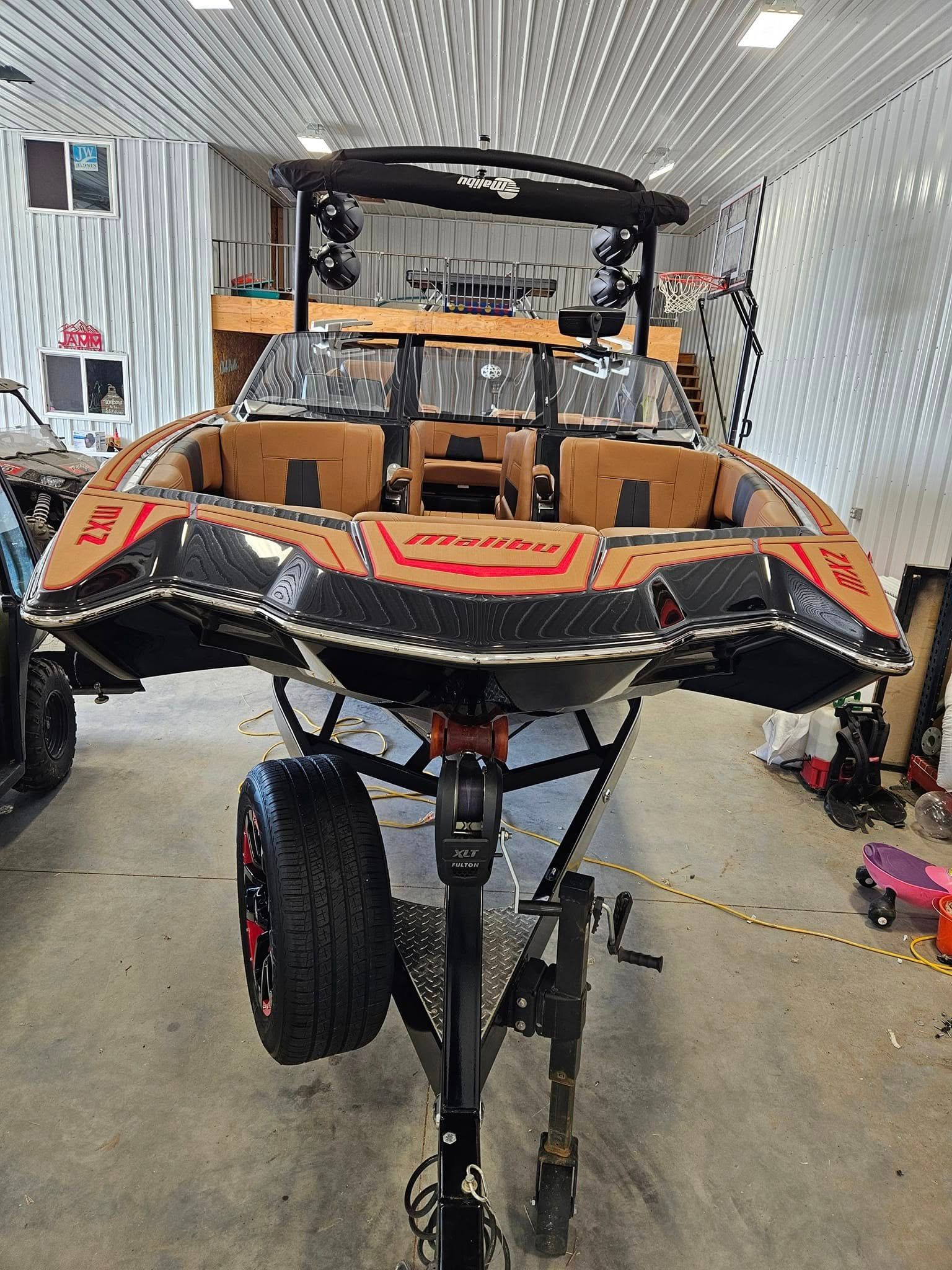 A black and tan wakeboard boat on a trailer inside a workshop, viewed from the rear.