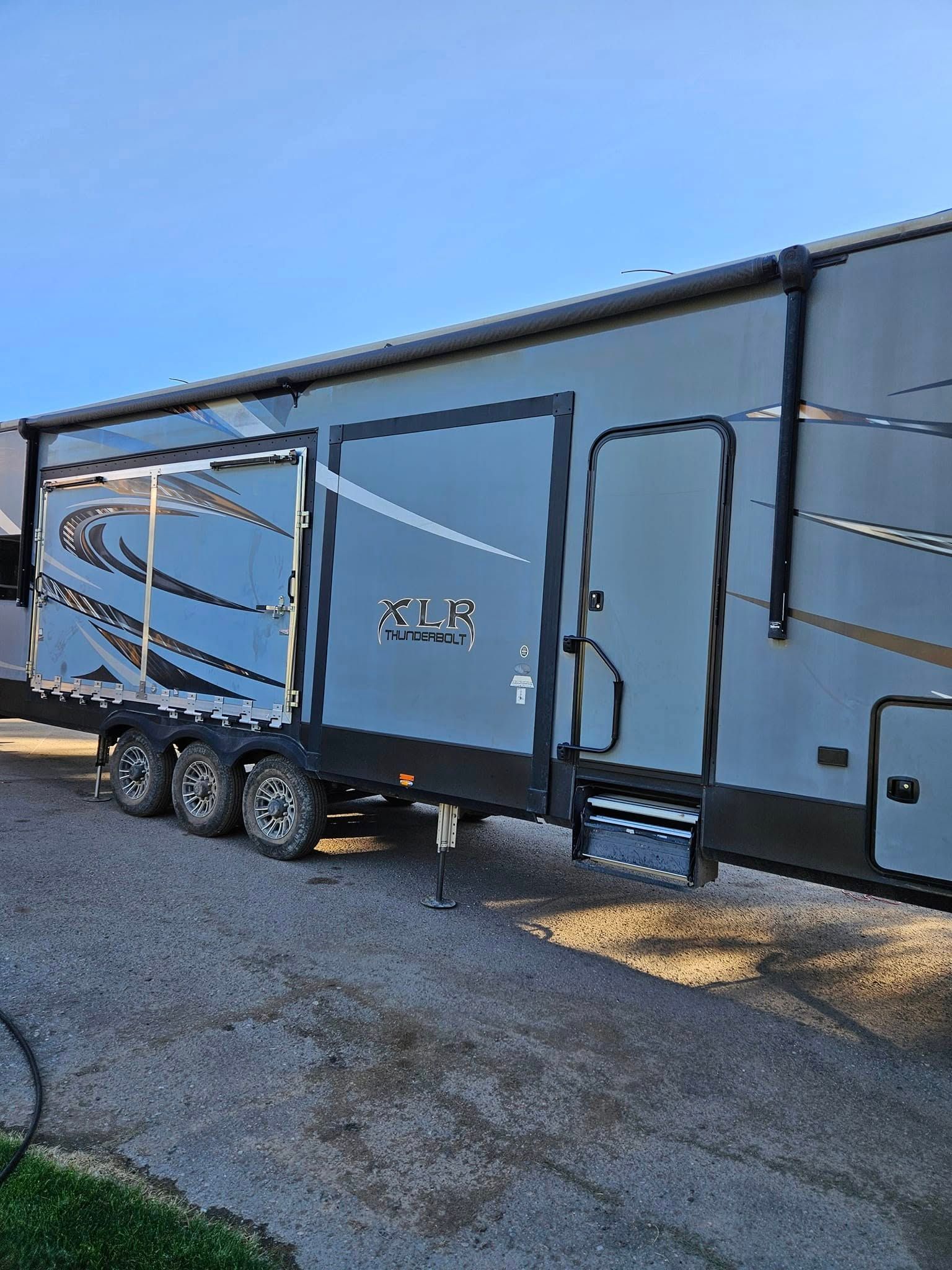 A grey XLR toy hauler RV parked on a gravel surface under a clear blue sky.