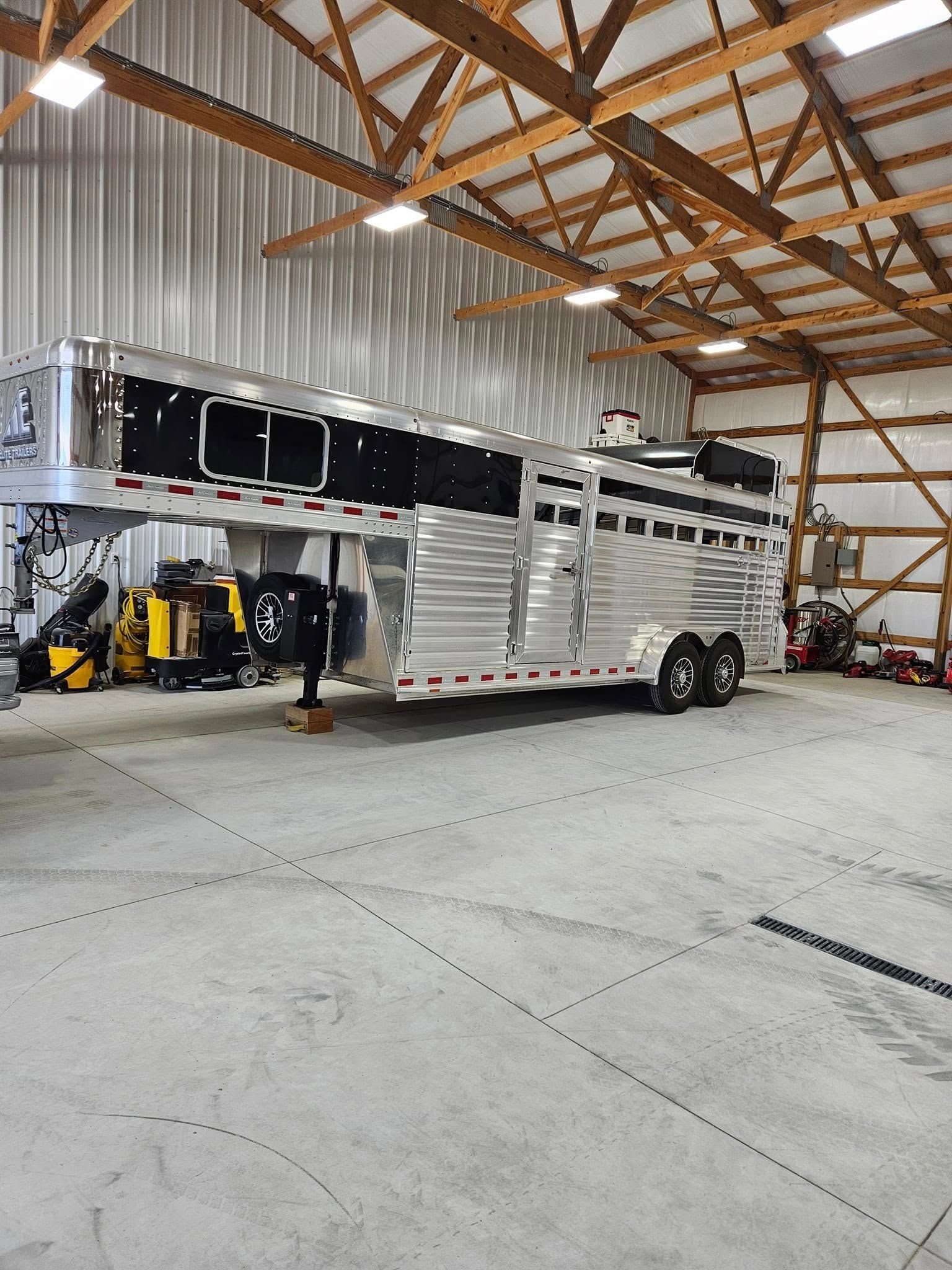 A silver and black gooseneck horse trailer parked inside a large, open metal-framed building with concrete flooring.