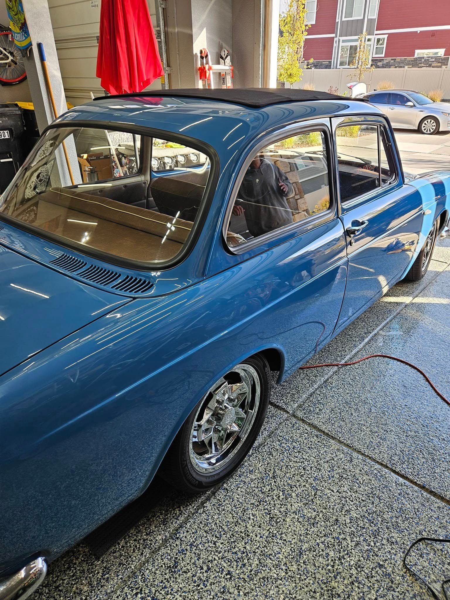 A blue vintage car with a black sunroof parked inside a garage on a speckled floor.