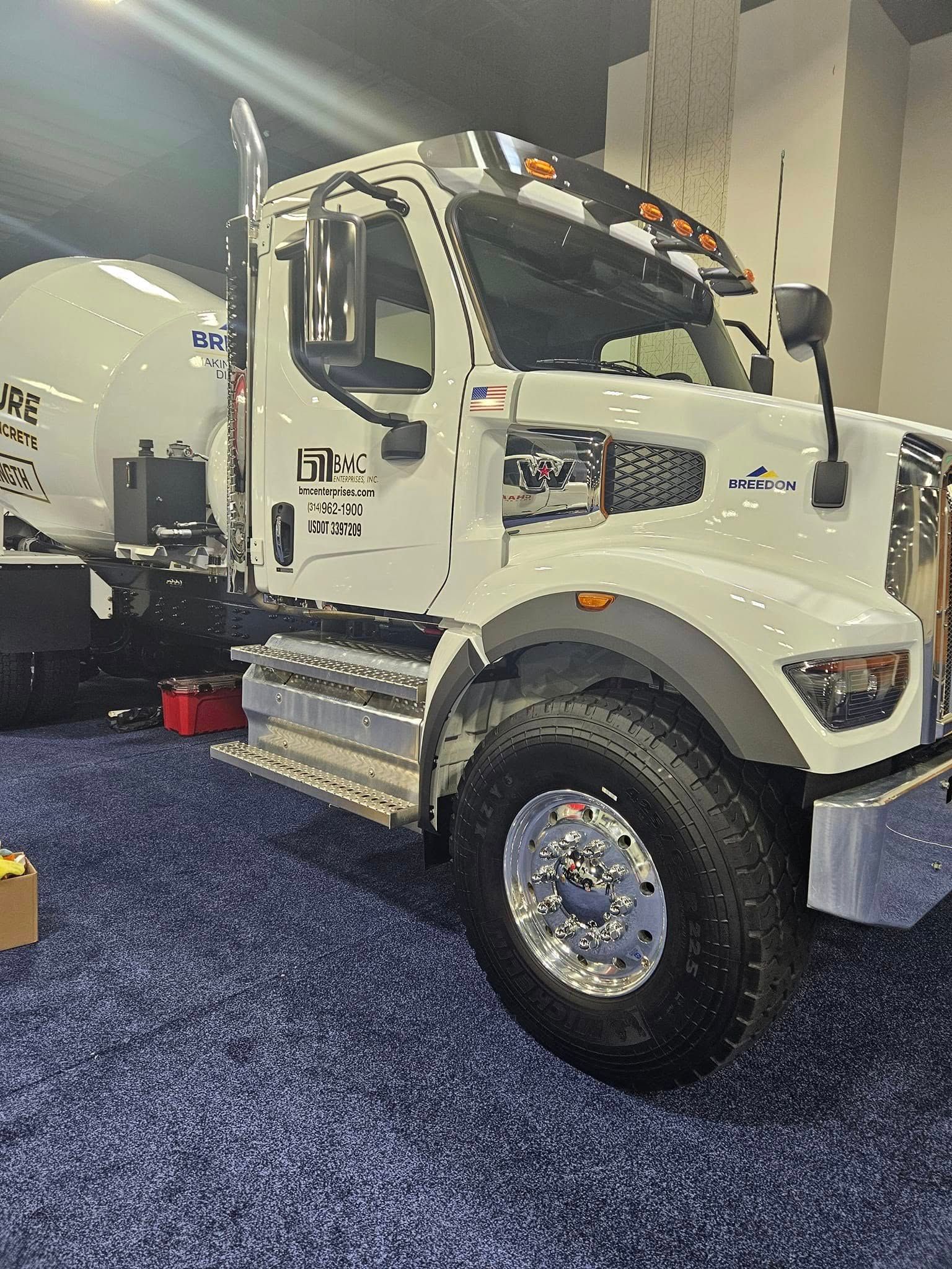 A white commercial cement mixer truck with chrome accents, parked on a blue carpeted indoor floor.